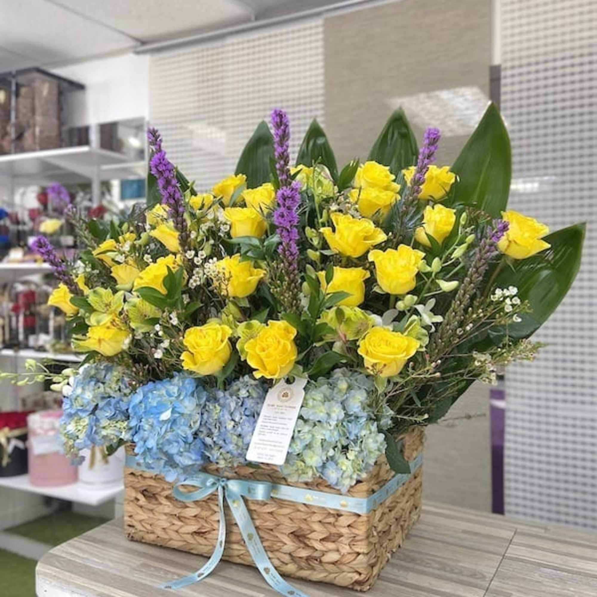 Large basket of yellow roses, blue hydrangeas, purple flowers and bold green leaves with a blue ribbon.