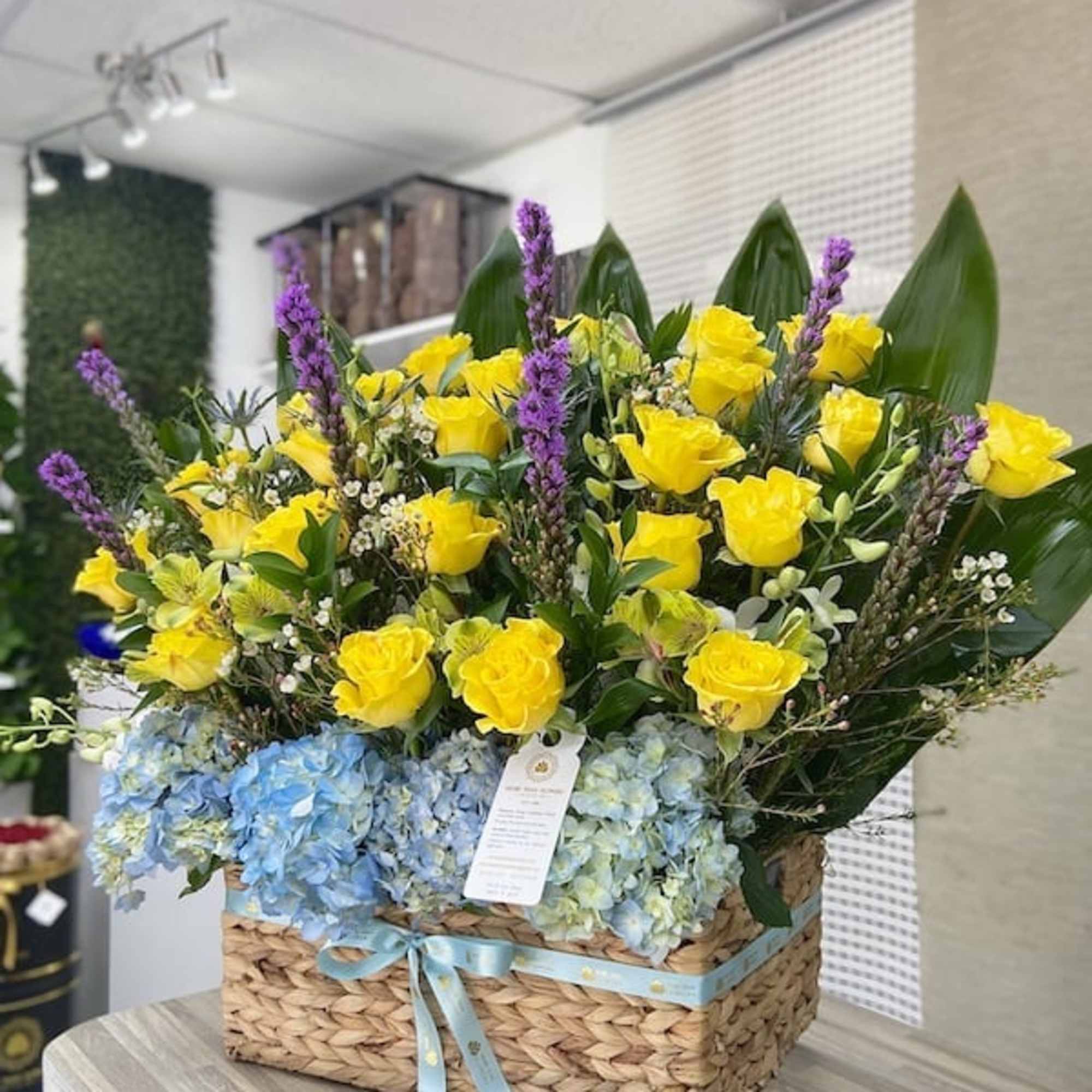 Large basket arrangement of yellow roses, blue hydrangeas, and tall purple flowers with a blue ribbon bow