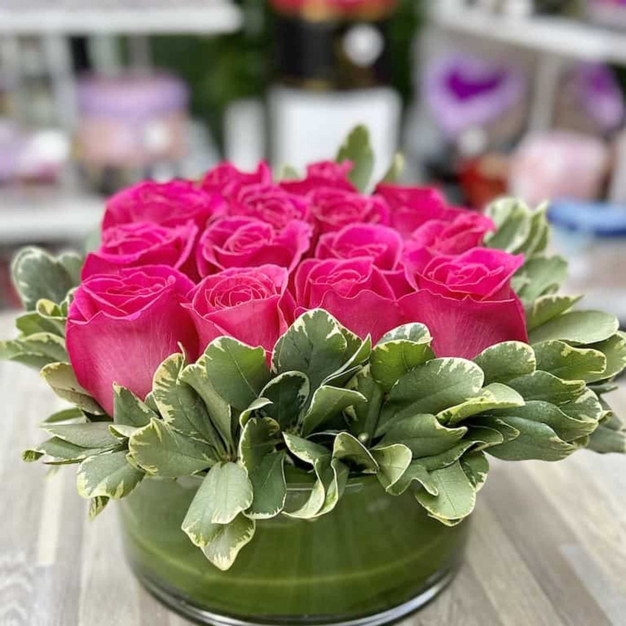Low arrangement of bright pink roses encircled by variegated green leaves in a clear glass vase.