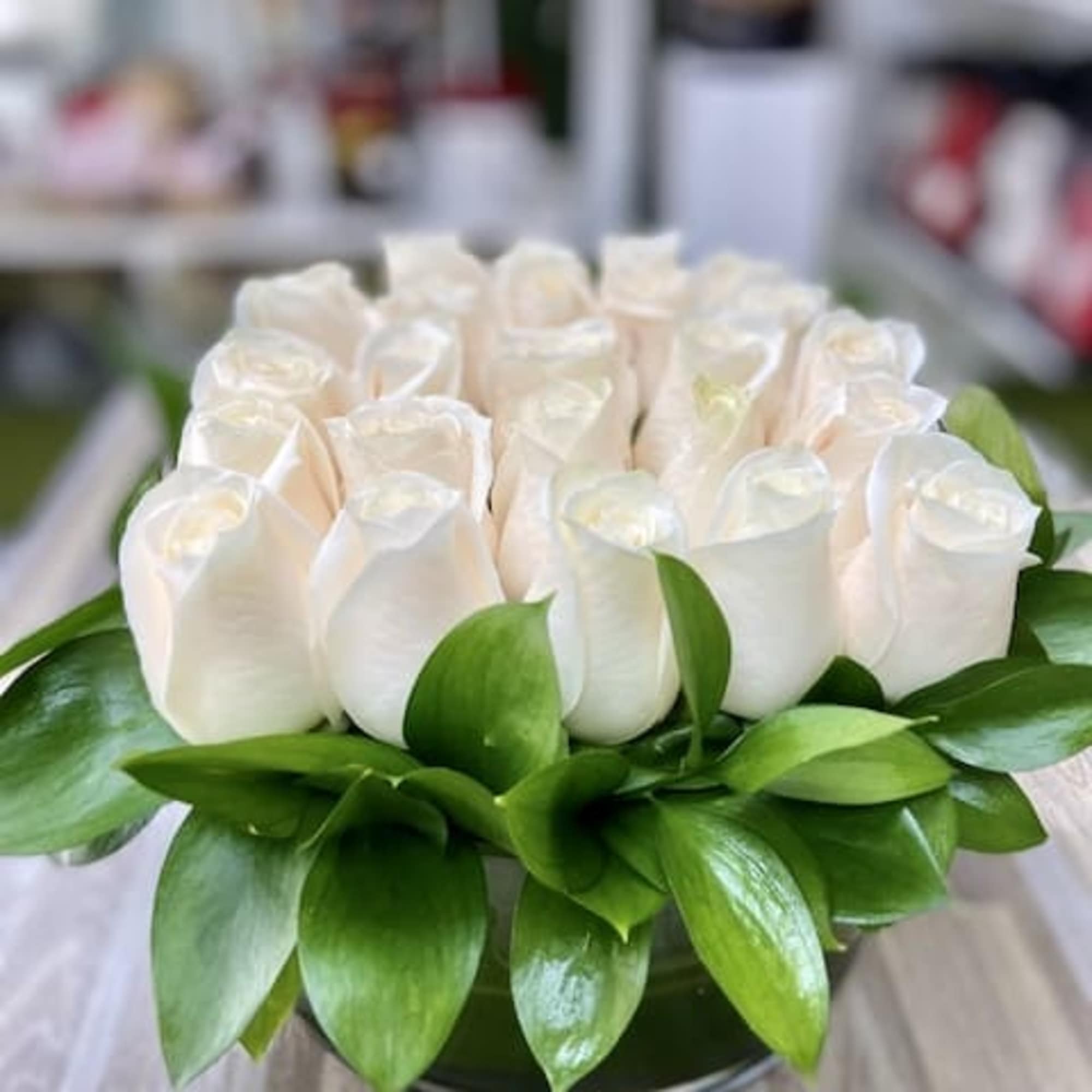 Low arrangement of cream roses tightly packed in a round glass vase with a collar of glossy green leaves.