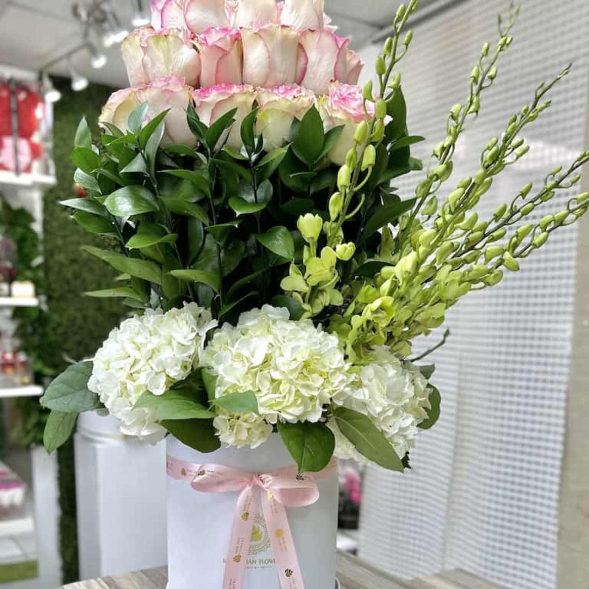 Tall arrangement of pink-edged roses, white hydrangeas, and green orchids in a white hat box with pink ribbon