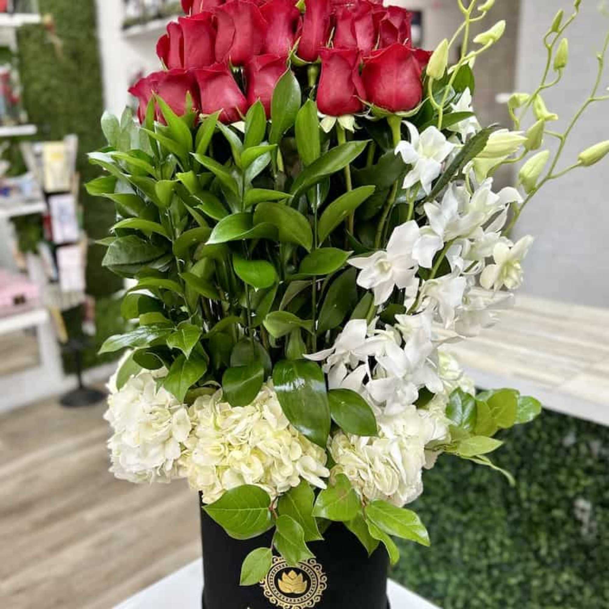 Tall arrangement of red roses, green foliage, and white flowers in a black hat box.