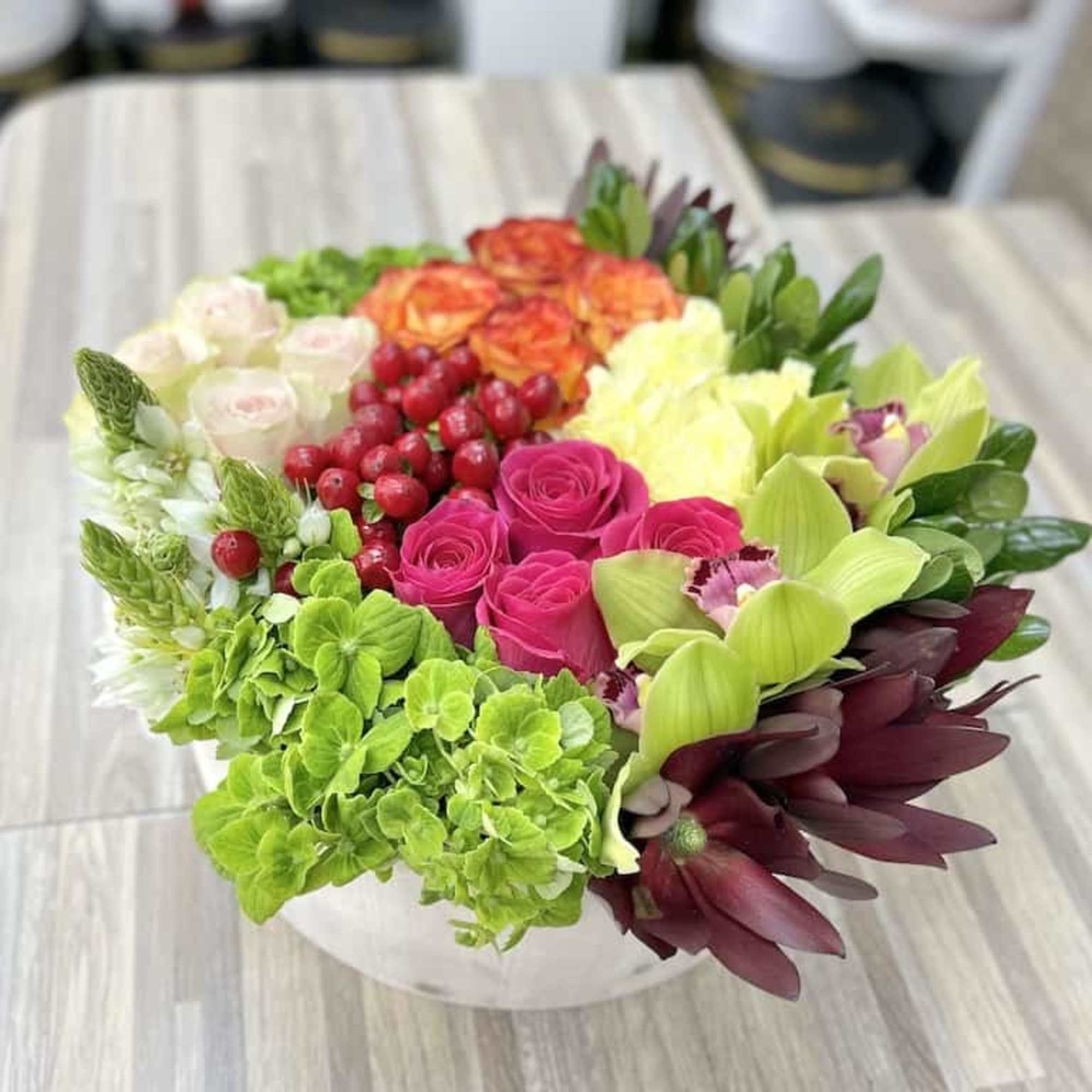 Low round centerpiece of bright roses, orchids, hydrangea, and mixed blooms in a white bowl.