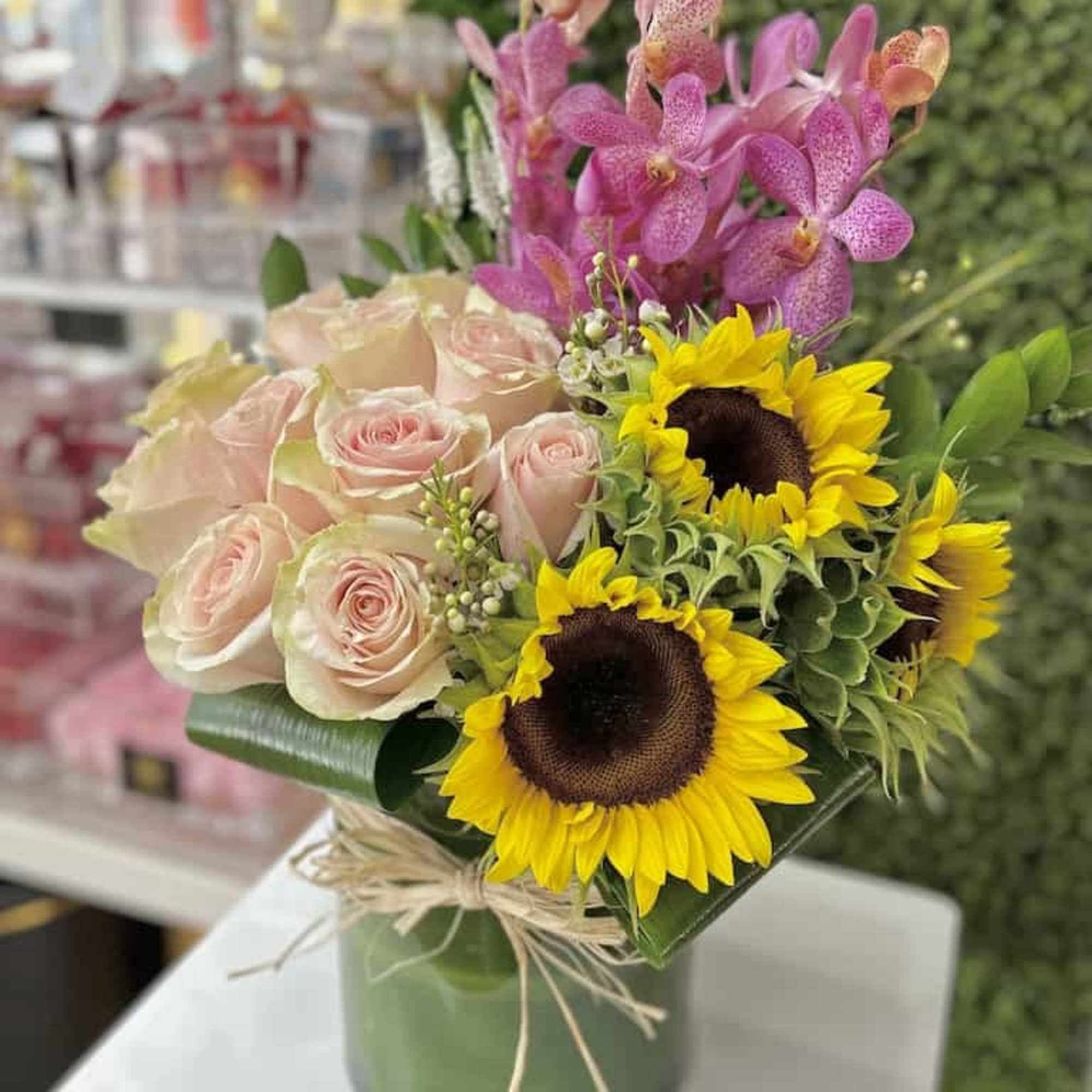 Arrangement of pale pink roses, yellow sunflowers, and pink orchids in a glass vase tied with raffia.