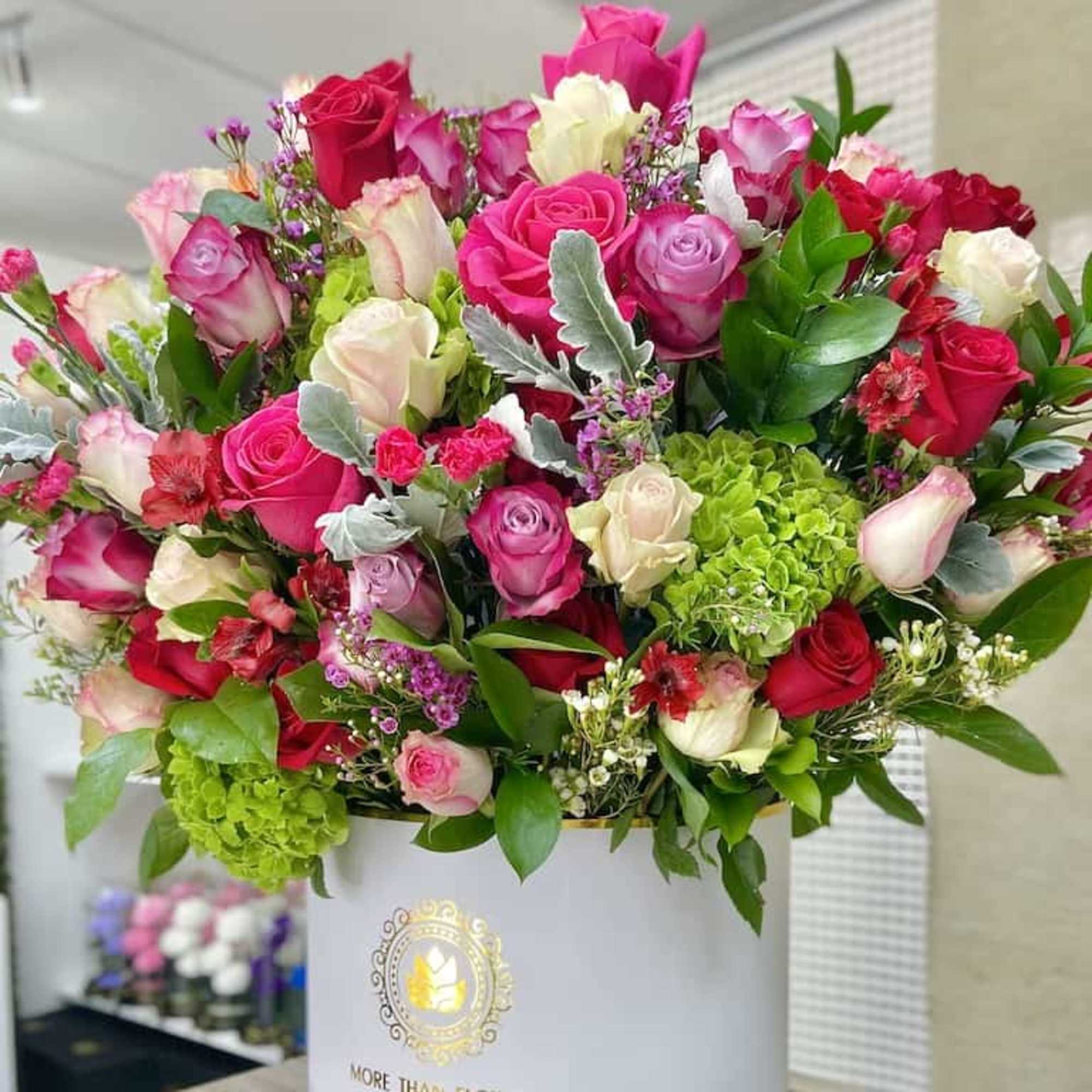 Large arrangement of pink, red, and cream roses with green hydrangea in a white hatbox container