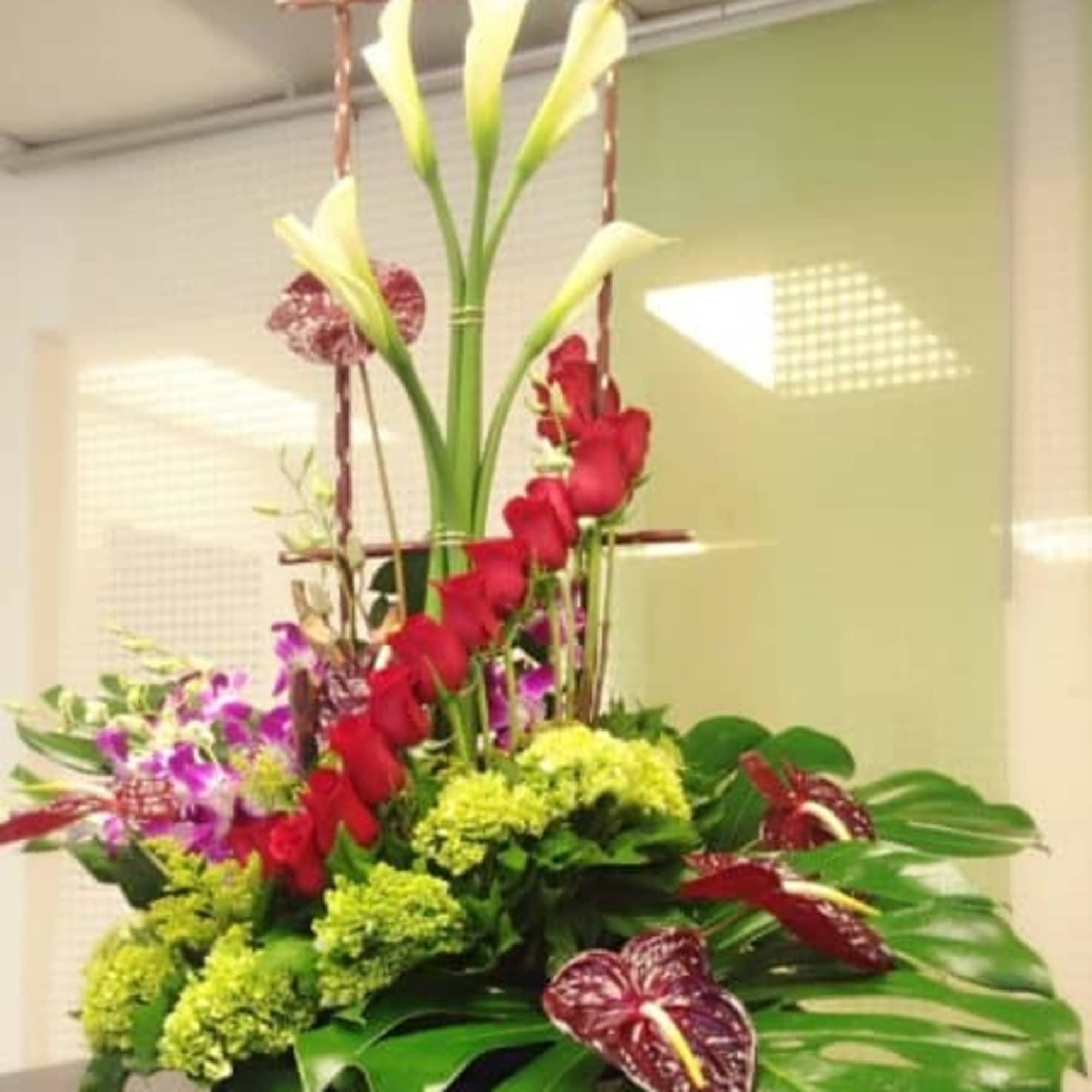 Tall arrangement with white calla lilies, red roses, green hydrangea, orchids and tropical leaves in a black container