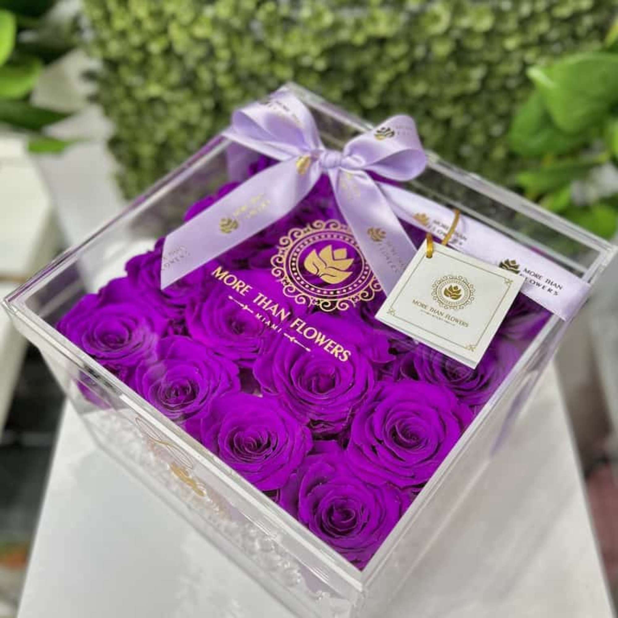 Square acrylic box filled with bright purple roses, topped with a lavender ribbon bow and gift tag