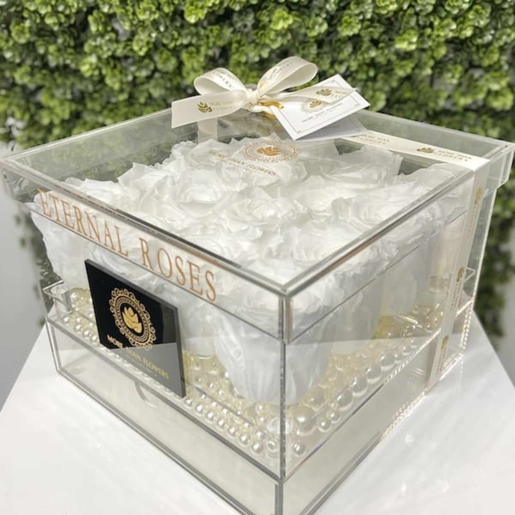 Clear acrylic box of white roses and pearls with ribboned lid against a leafy backdrop