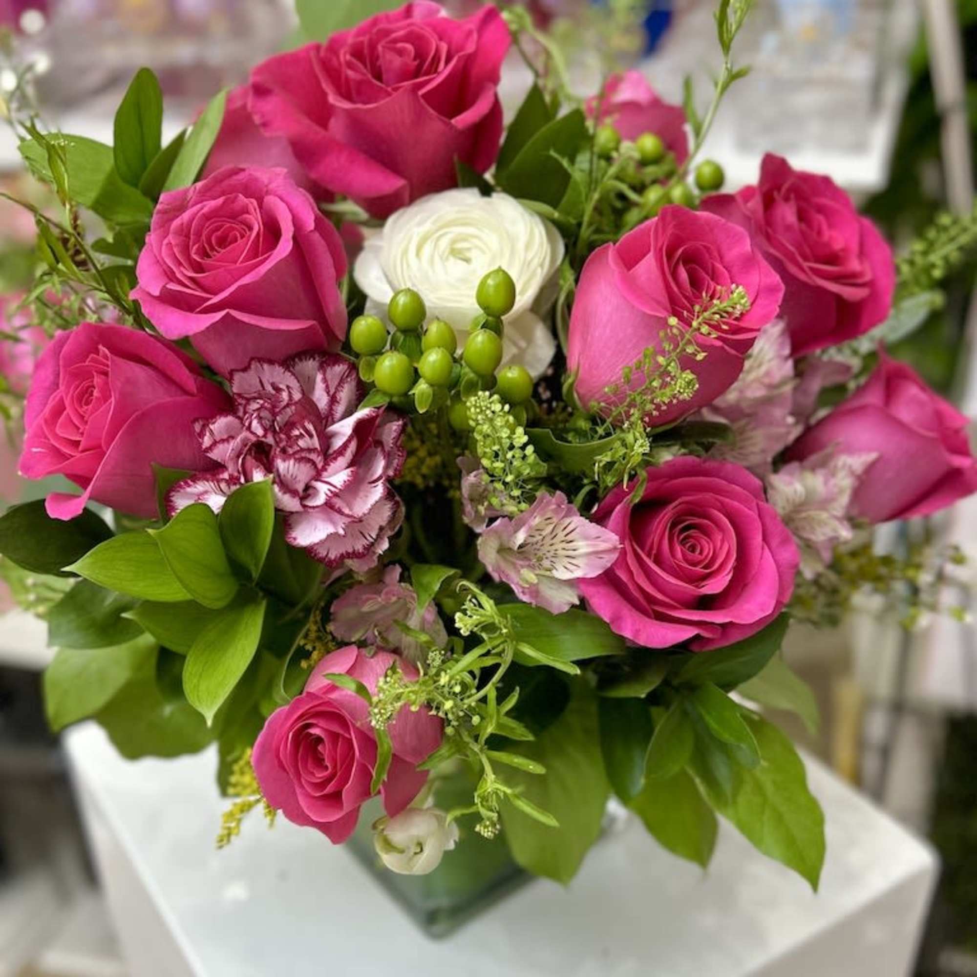 Bright pink rose arrangement with white accent blooms and green berries in a clear glass cube vase