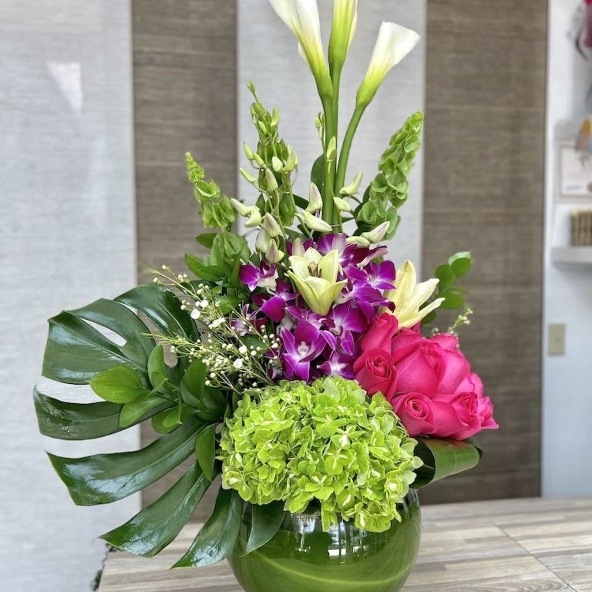 Tall arrangement of white calla lilies, pink roses, orchids, and green hydrangea in a green glass vase