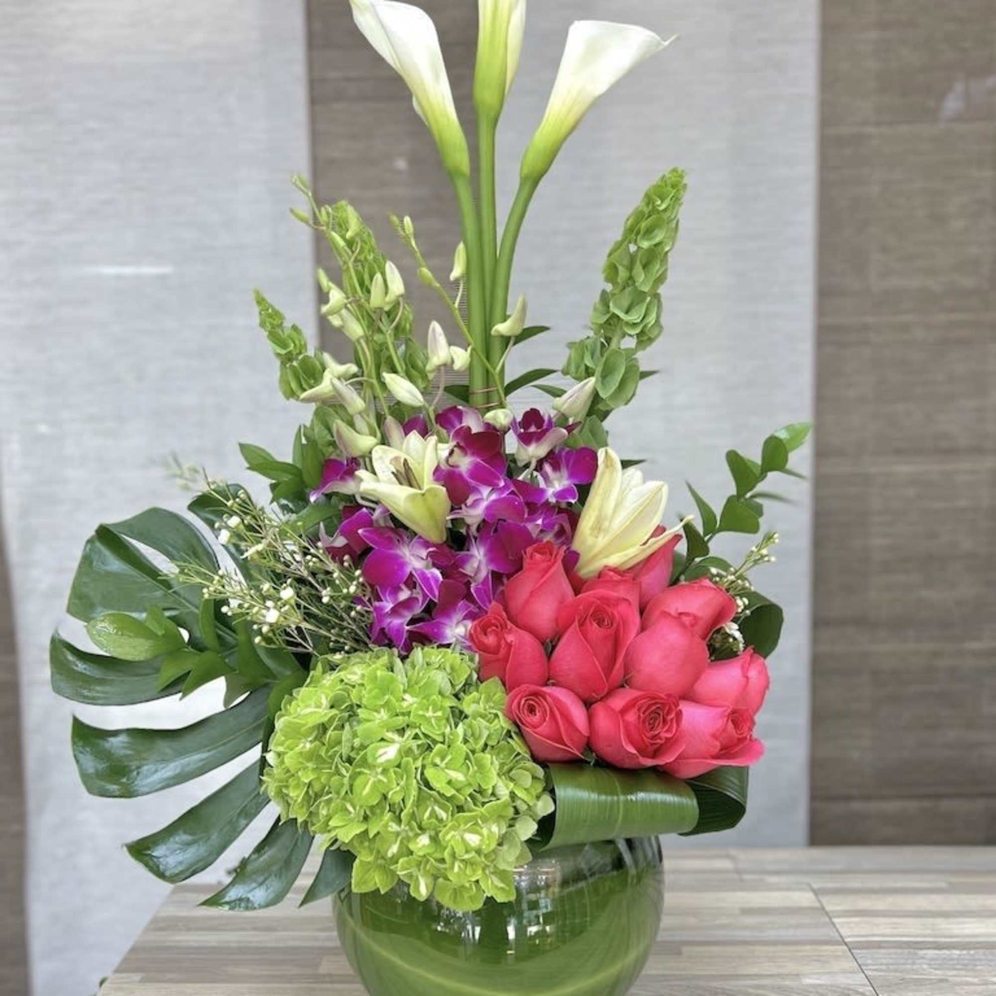 Tall arrangement of white calla lilies, pink roses, orchids, and green hydrangea in a round glass vase