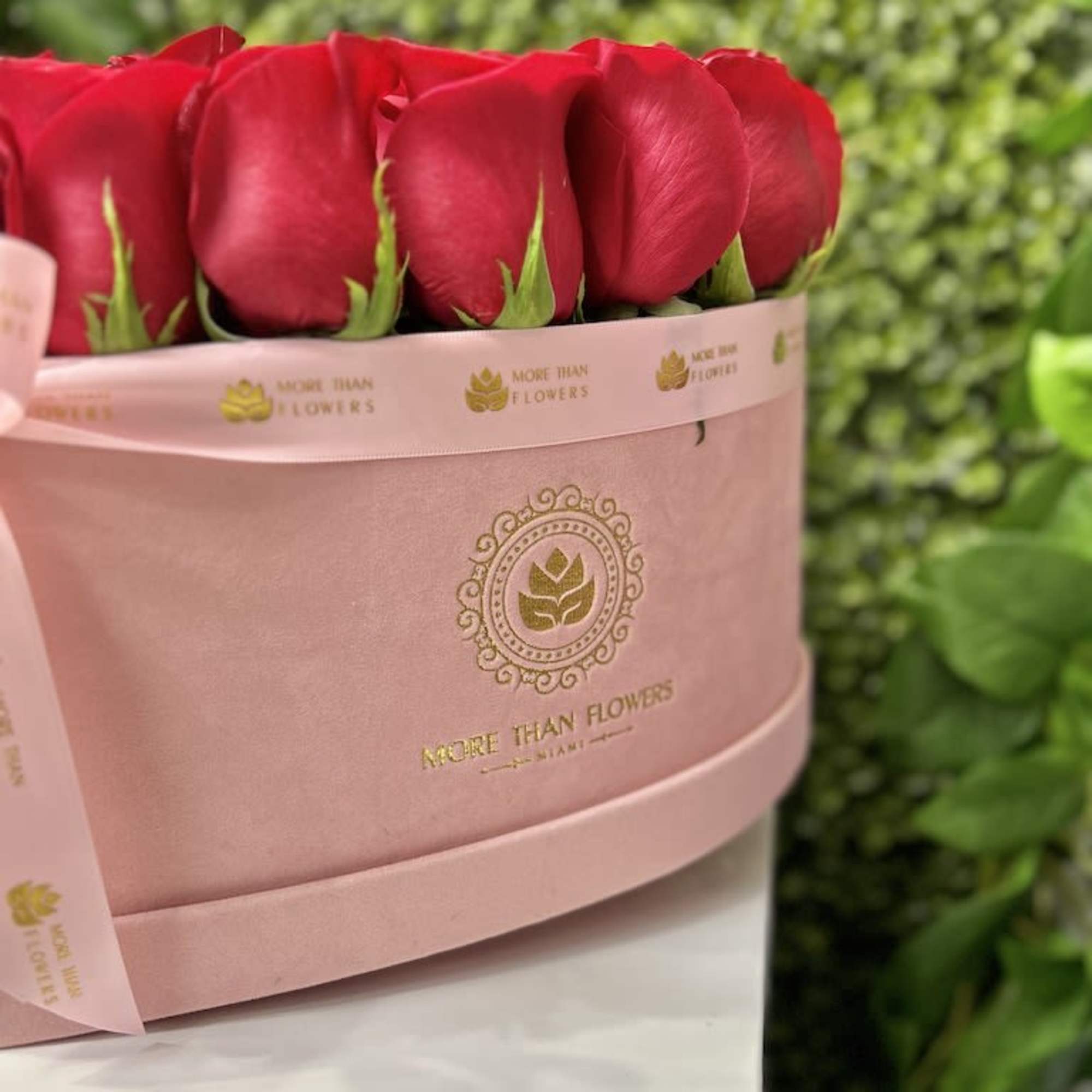Round pink hatbox filled with upright red roses and wrapped with a branded pink ribbon.
