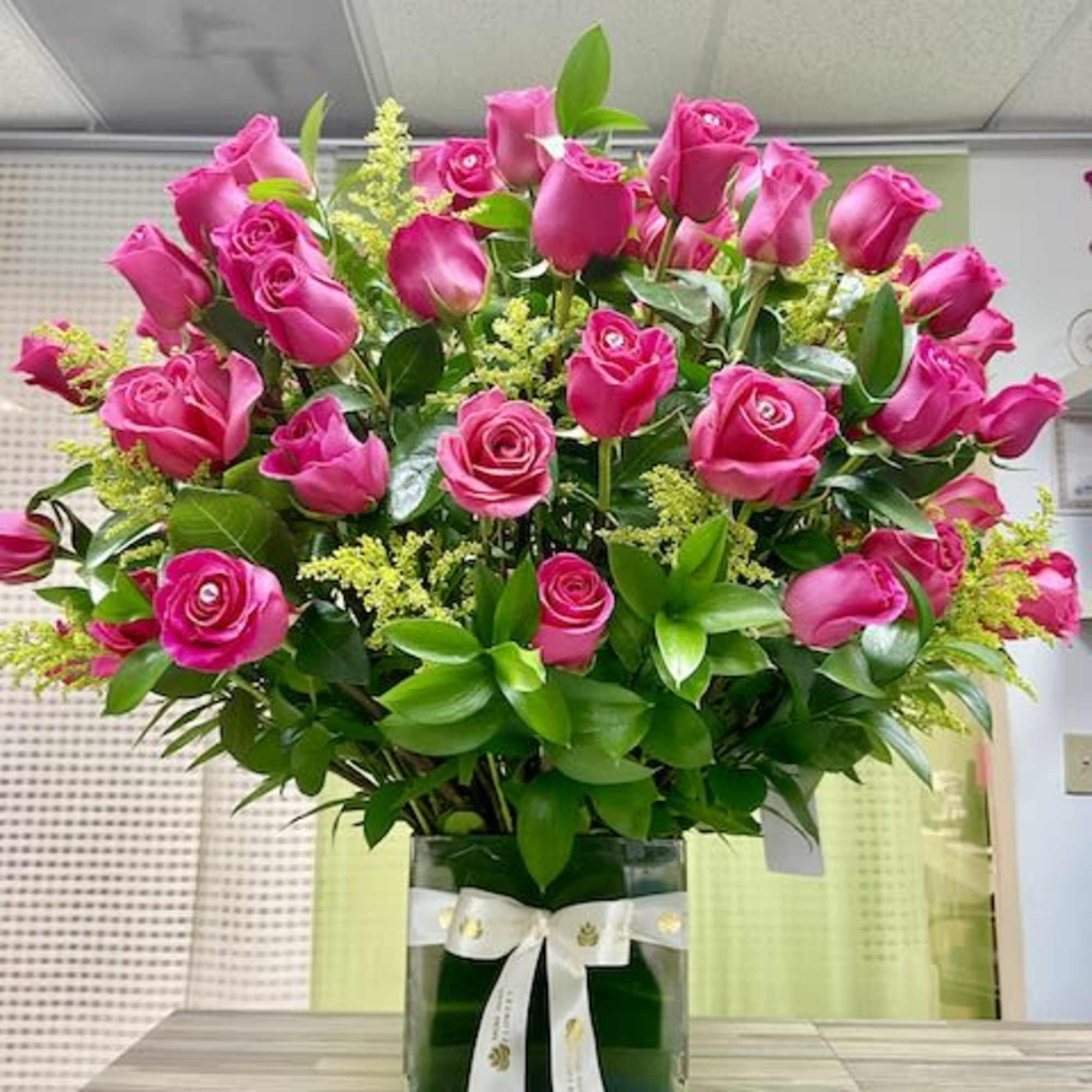 Tall arrangement of bright pink roses with filler flowers in a square glass vase with a white ribbon