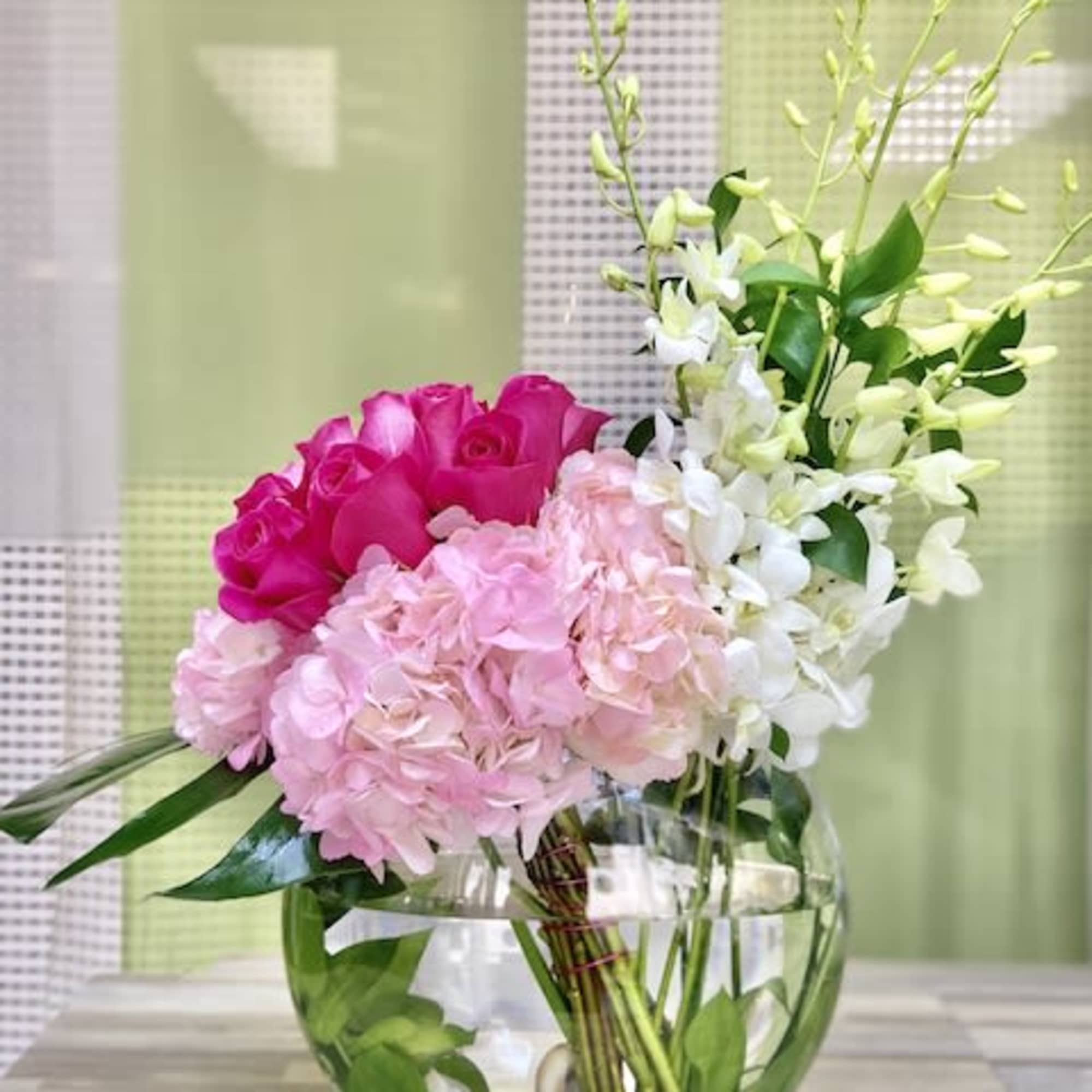 Pink roses, hydrangeas, and white orchids arranged in a round glass vase with stones.
