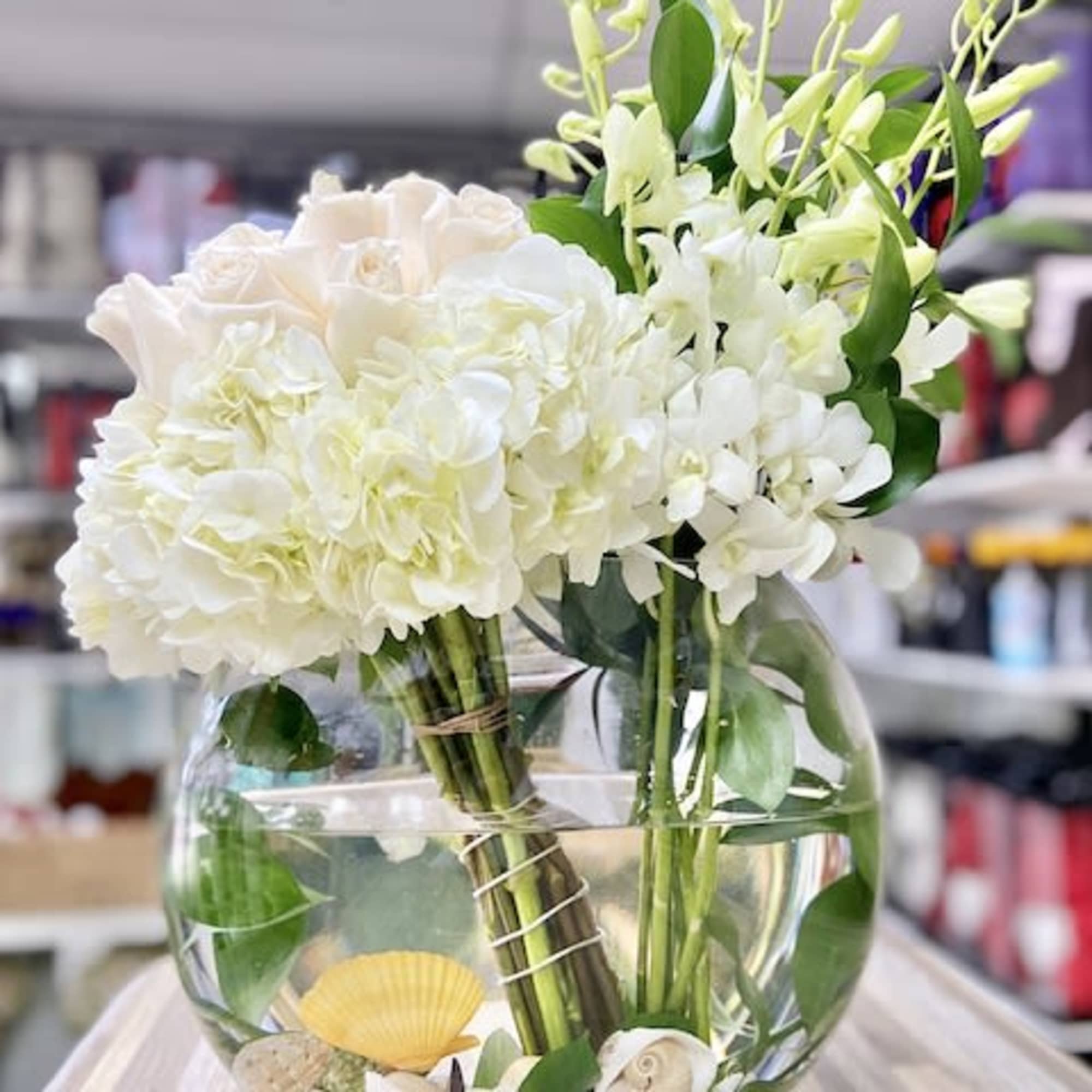 Round glass vase with white hydrangeas, pale roses, and orchids over seashells in water