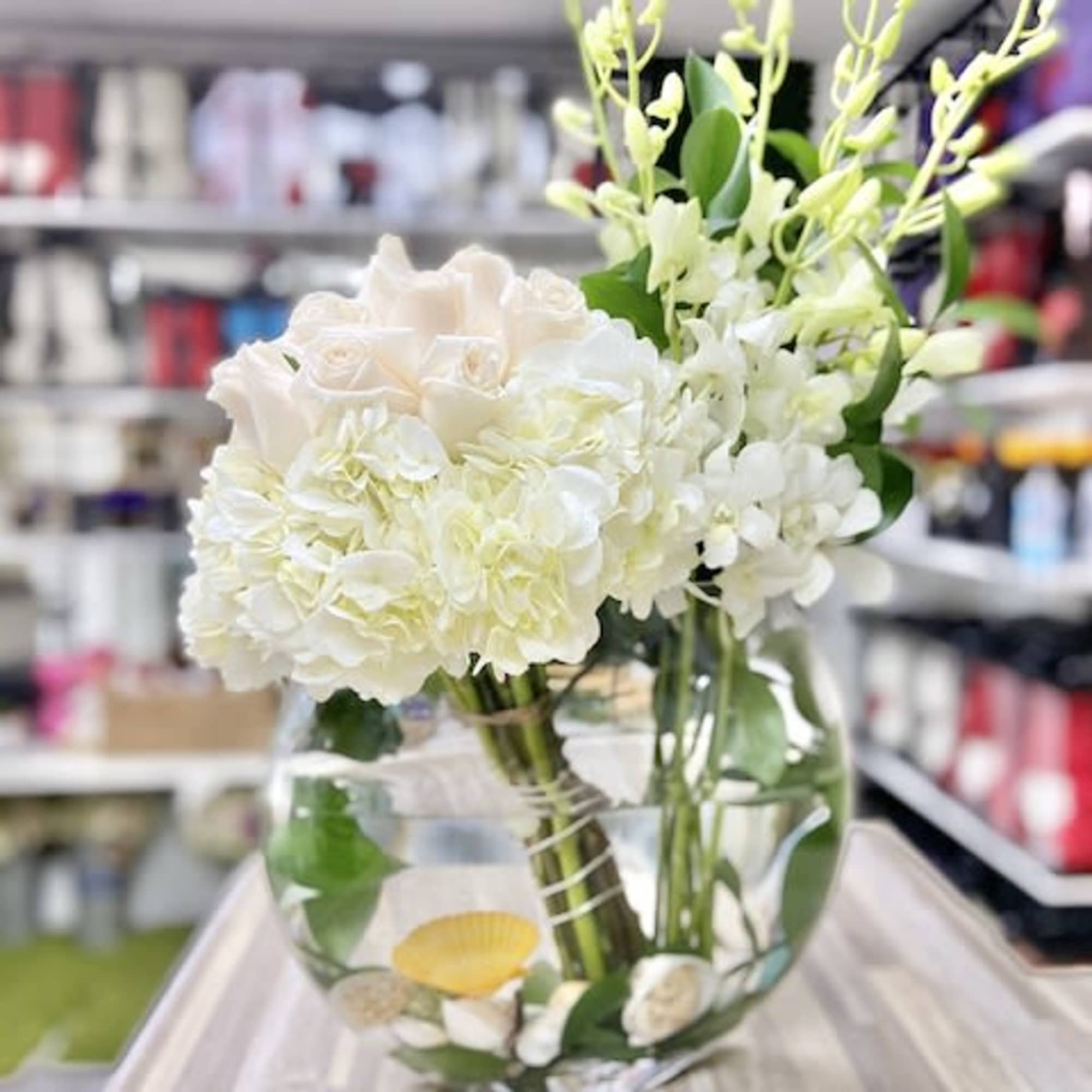 Round glass vase of white roses, hydrangeas, and orchids with seashells in the water