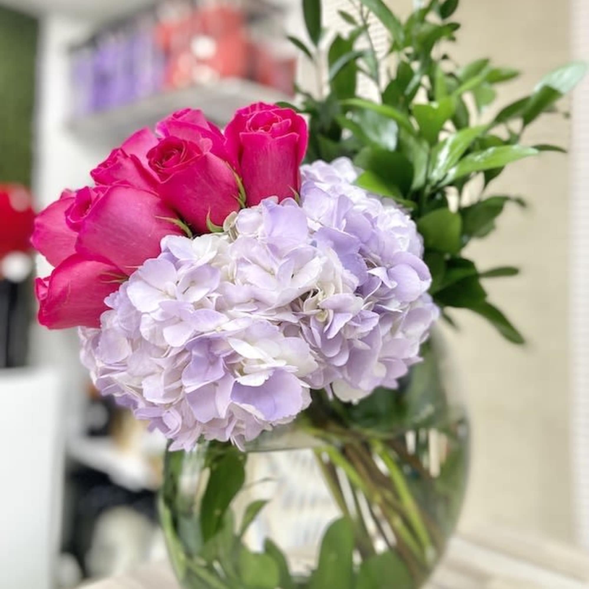 Round glass vase with pink roses, pale lavender hydrangeas, and tall greenery arranged to one side.
