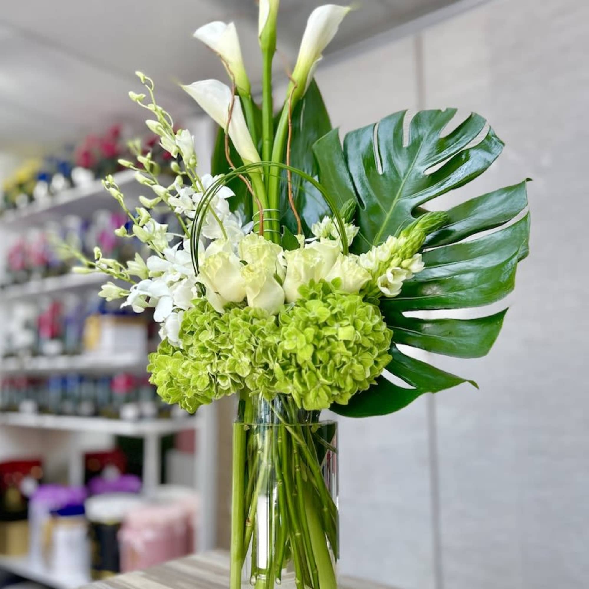Tall arrangement of white calla lilies, orchids, and green hydrangeas with a large tropical leaf in a clear glass vase