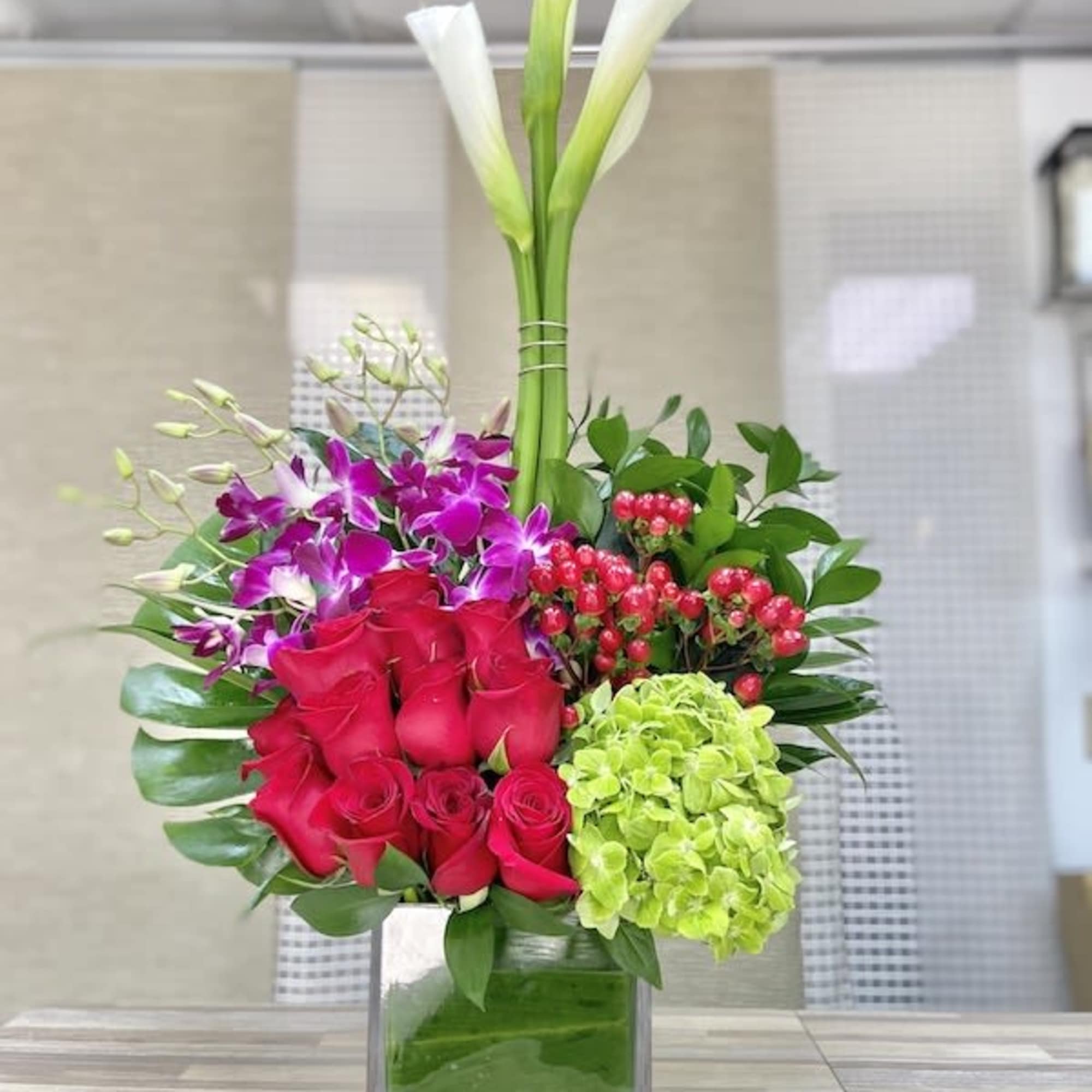 Tall modern arrangement with white calla lilies, red roses, purple orchids, and green hydrangea in a glass cube vase.