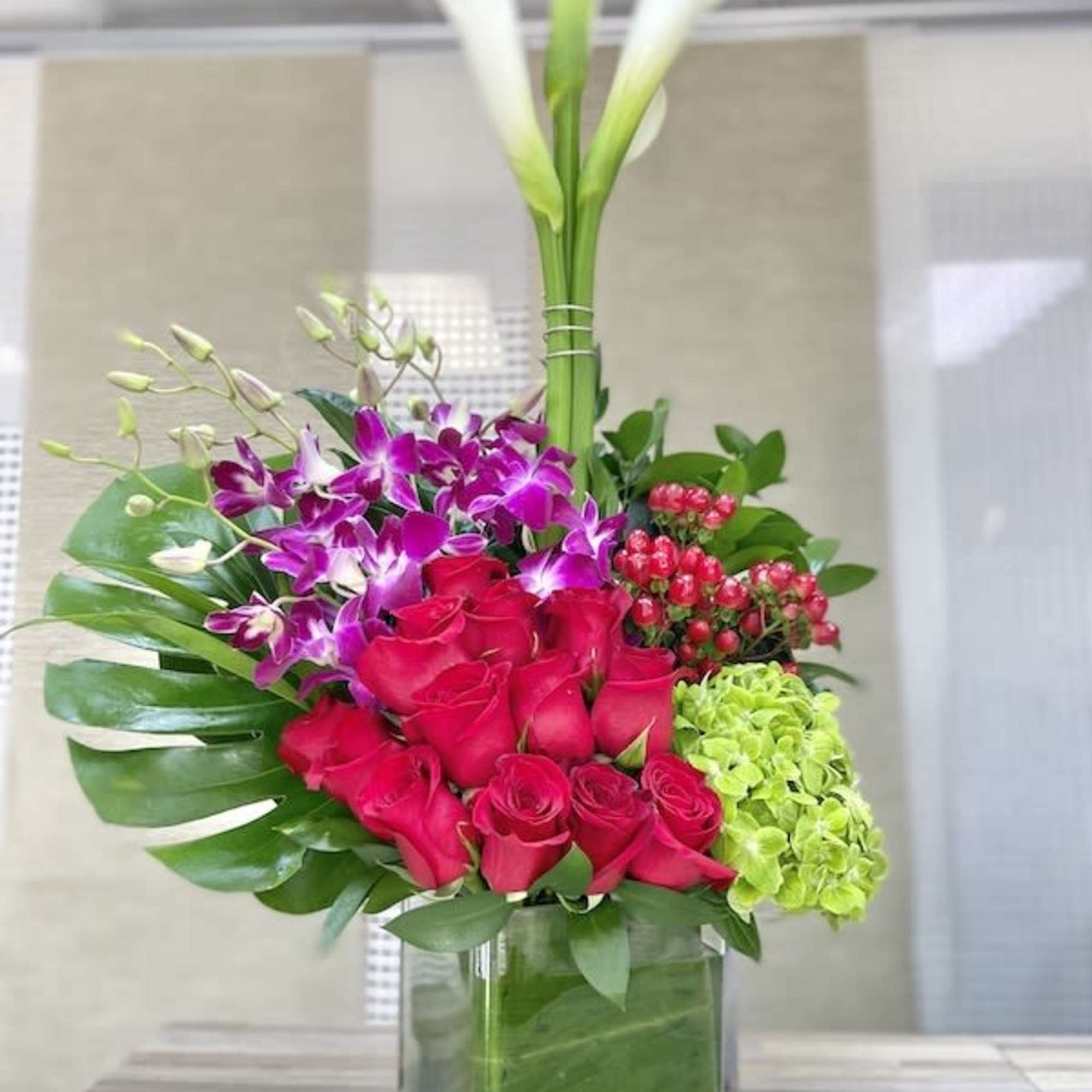 Tall mixed arrangement with red roses, purple orchids, green hydrangea, and white calla lilies in a glass cube vase