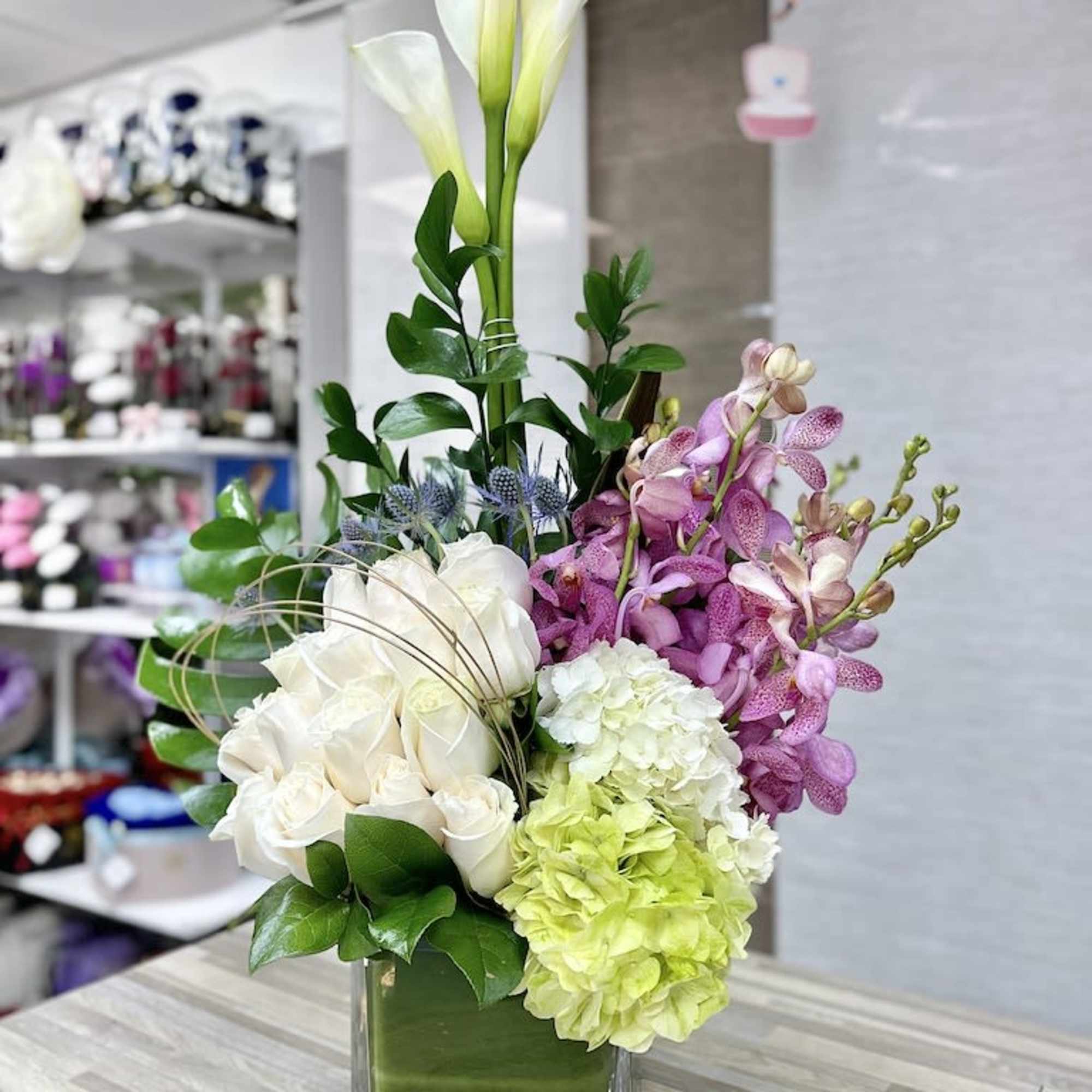 Tall mixed arrangement with white calla lilies, roses, hydrangeas and pink orchids in a clear square vase