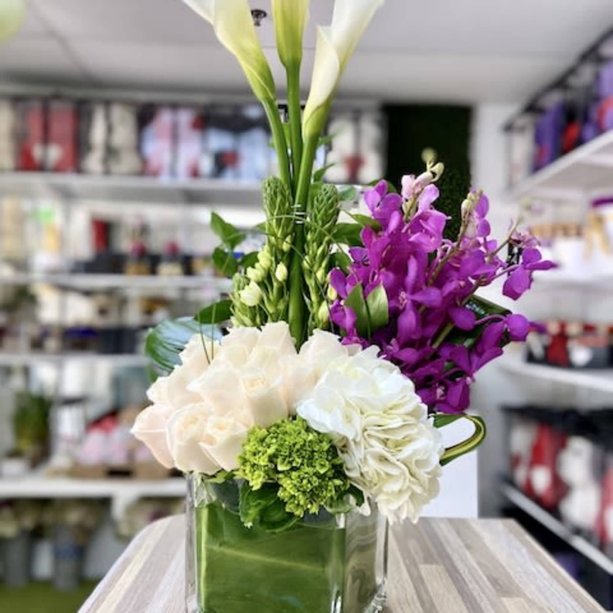 Tall white calla lilies with purple orchids and pale roses in a green-lined glass cube vase.