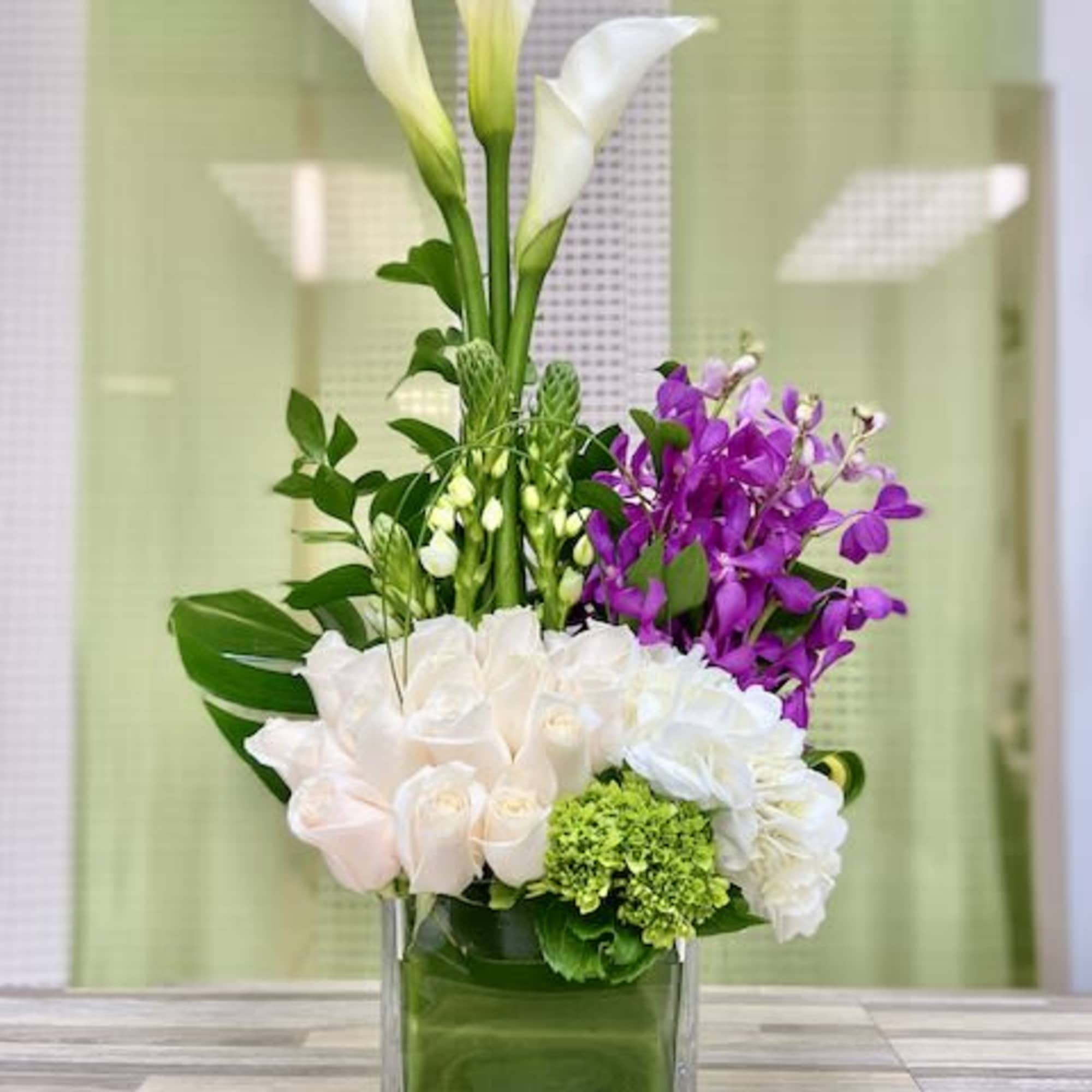 Tall modern arrangement with white calla lilies, cream roses, and purple orchids in a glass cube vase