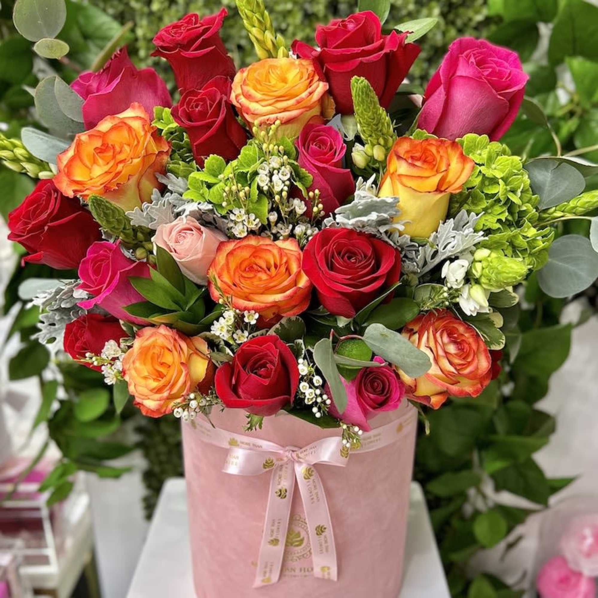 Bright mix of red, pink, and orange roses arranged in a pink hat box with ribbon