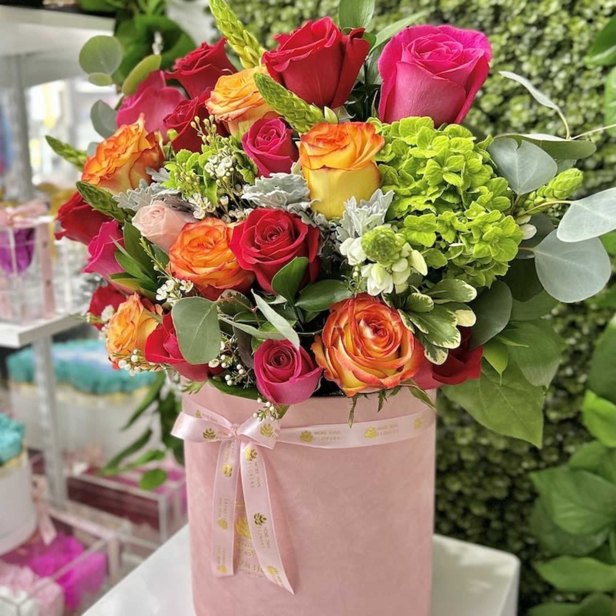 Bright arrangement of pink, red, and orange roses with green hydrangea in a pink hat box.