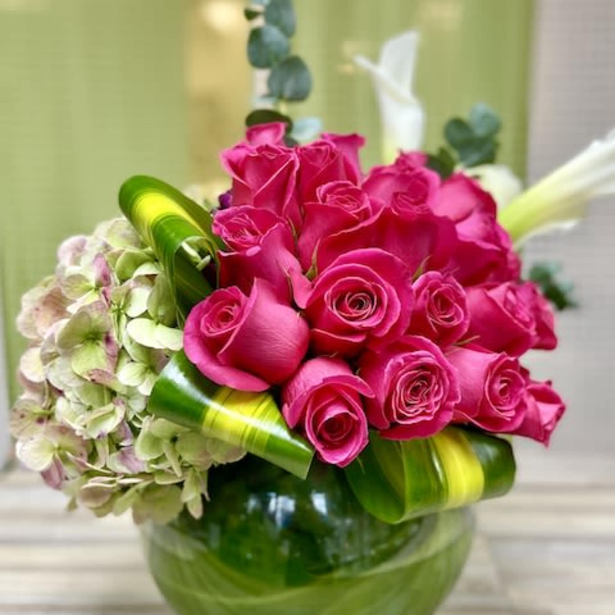 Low glass vase with bright pink roses, pale hydrangea, and white calla lilies arranged with bold folded leaves.
