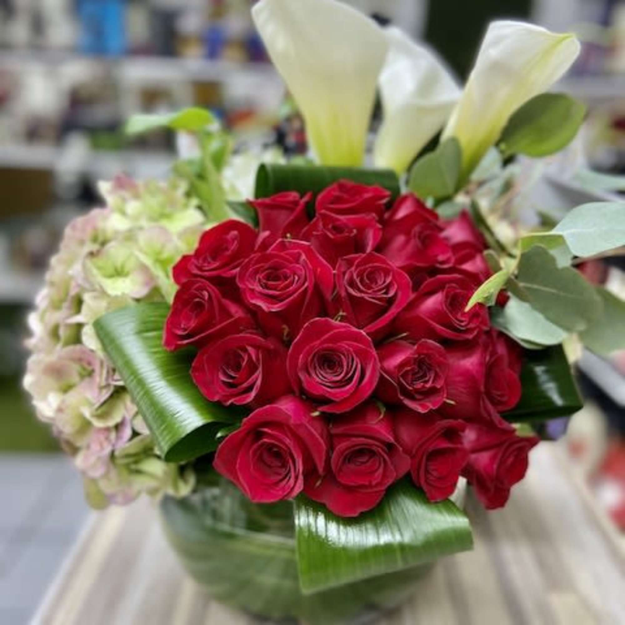 Large cluster of red roses with white calla lilies and hydrangea in a clear glass bowl with folded green leaves.
