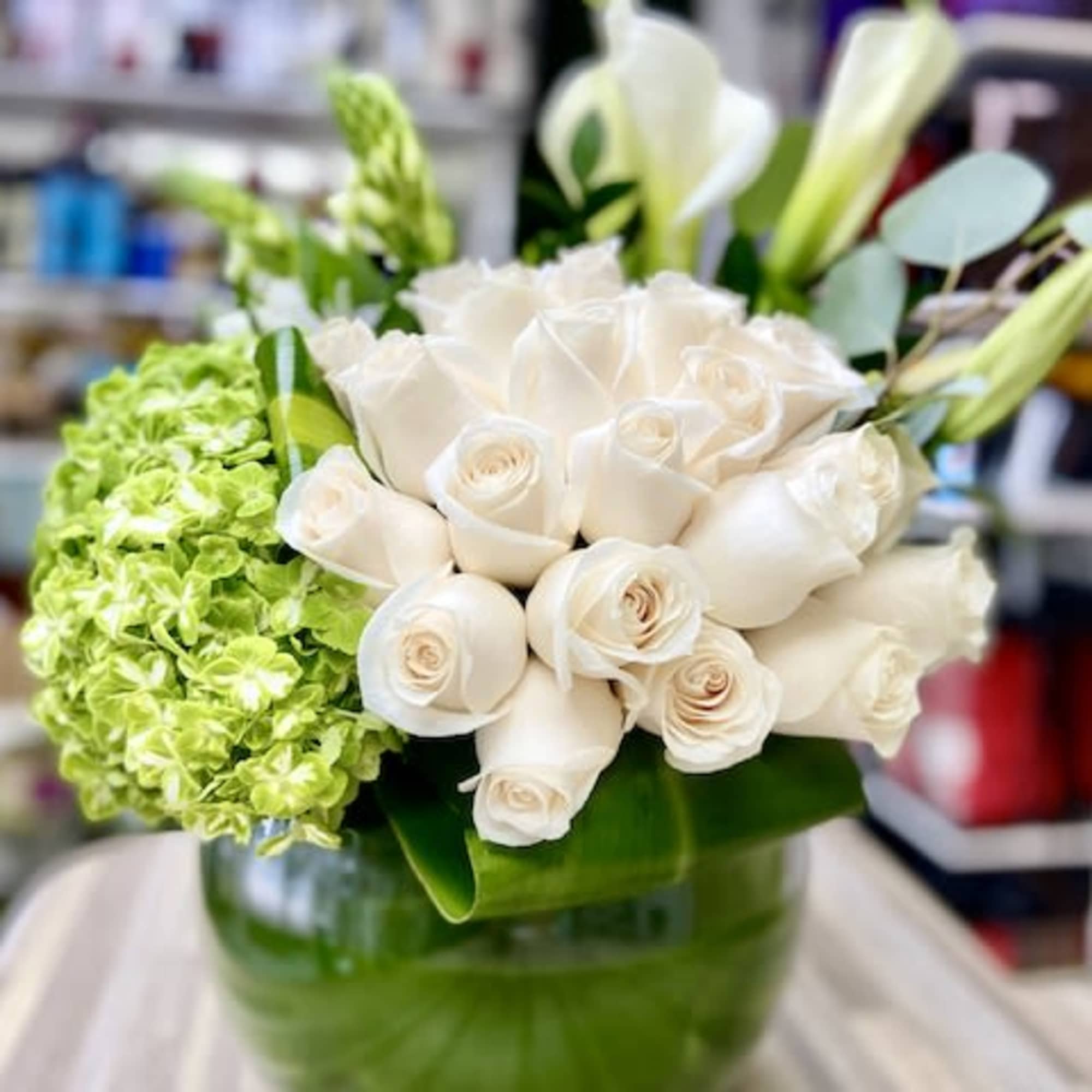 Round glass vase filled with white roses, white calla lilies, and green hydrangeas.