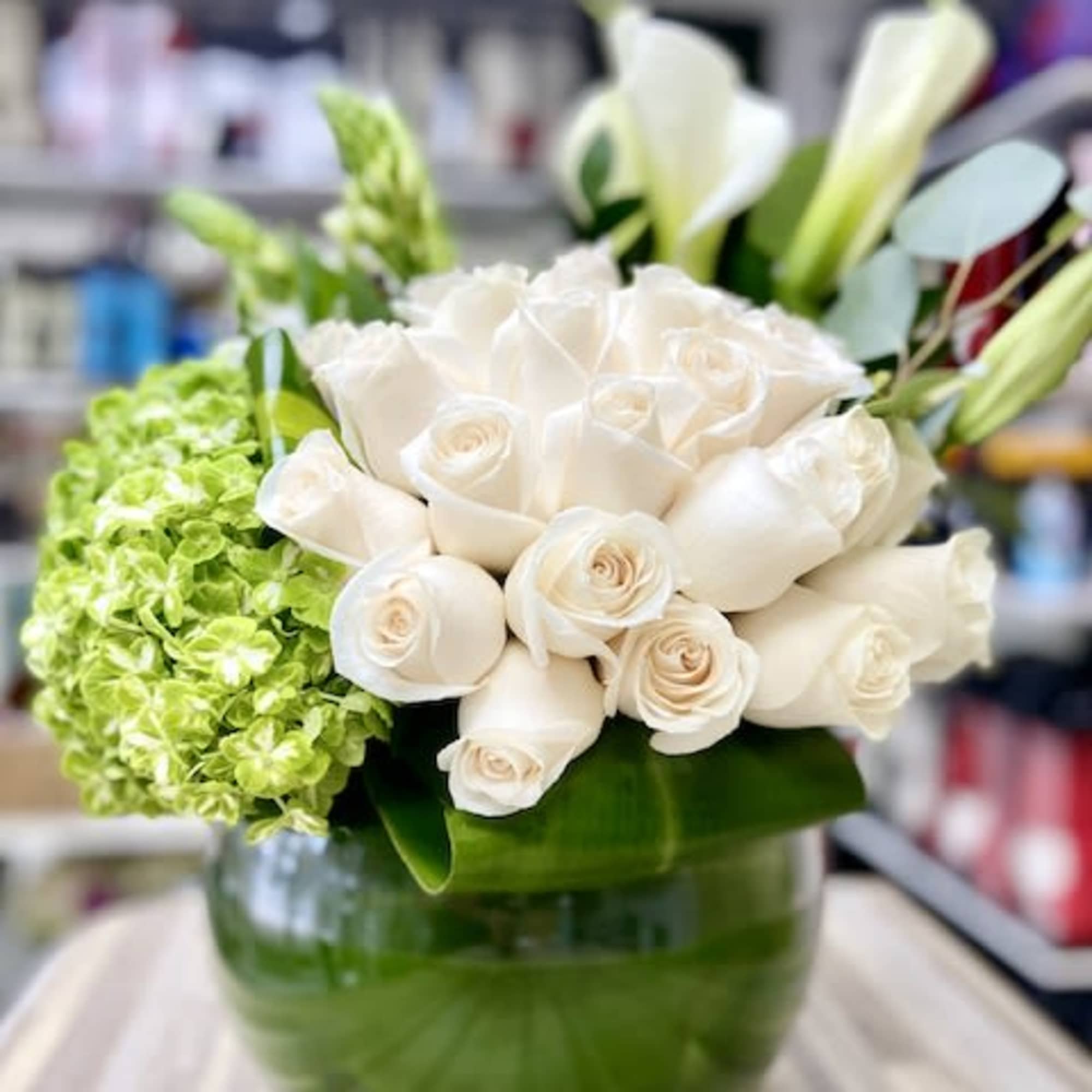 Low round arrangement of ivory roses, green hydrangea, and white calla lilies in a glass bowl vase.