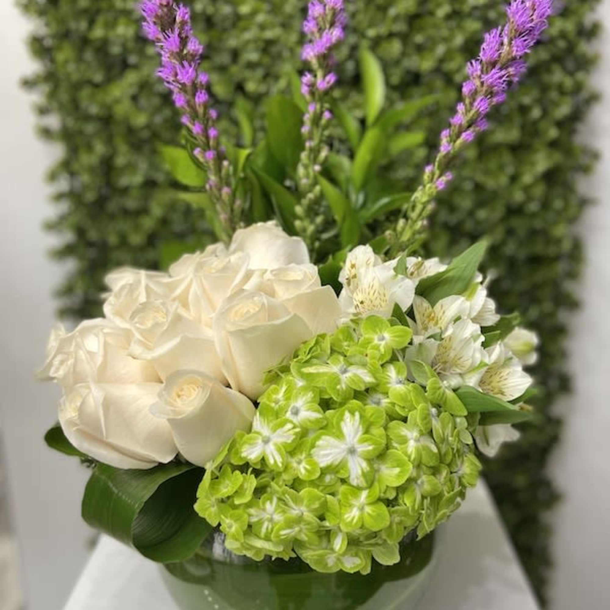 Compact arrangement of white roses, lime hydrangea, and purple spikes in a green bowl vase