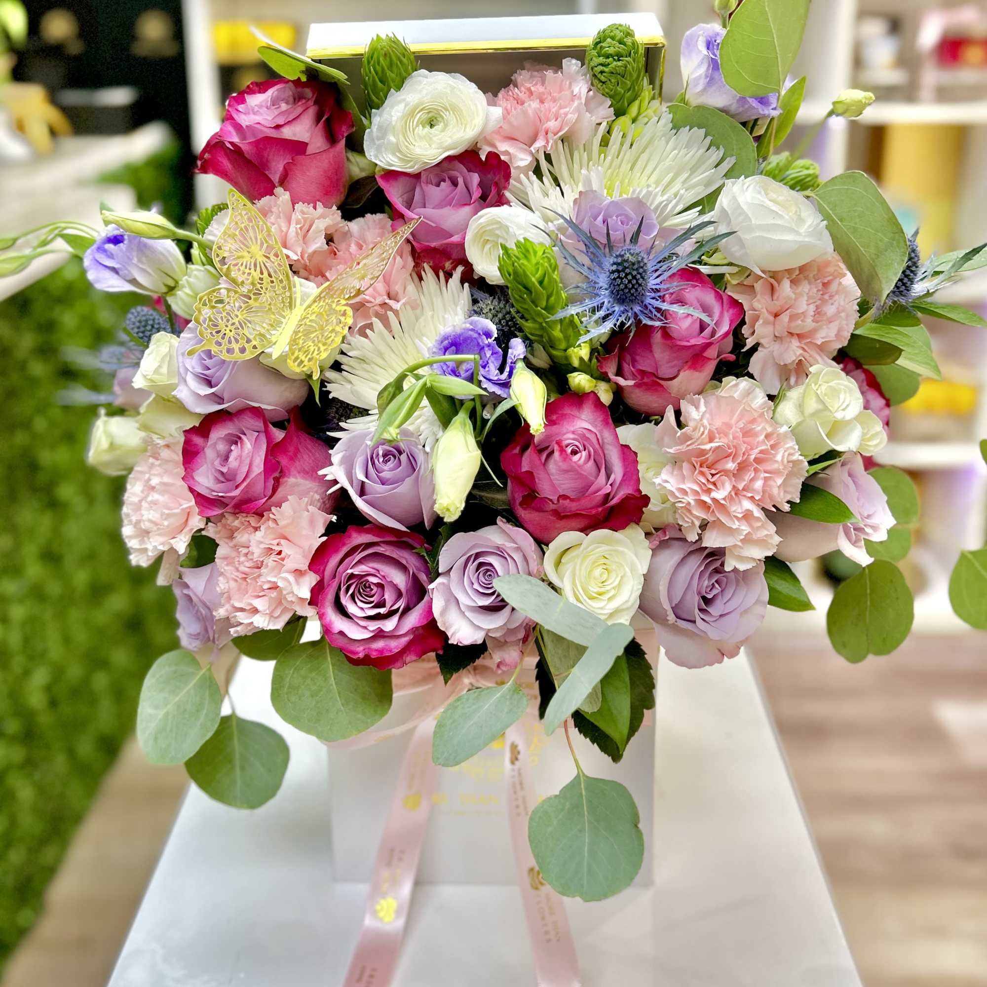 Boxed arrangement of pink, white, and lavender flowers with a decorative gold butterfly.