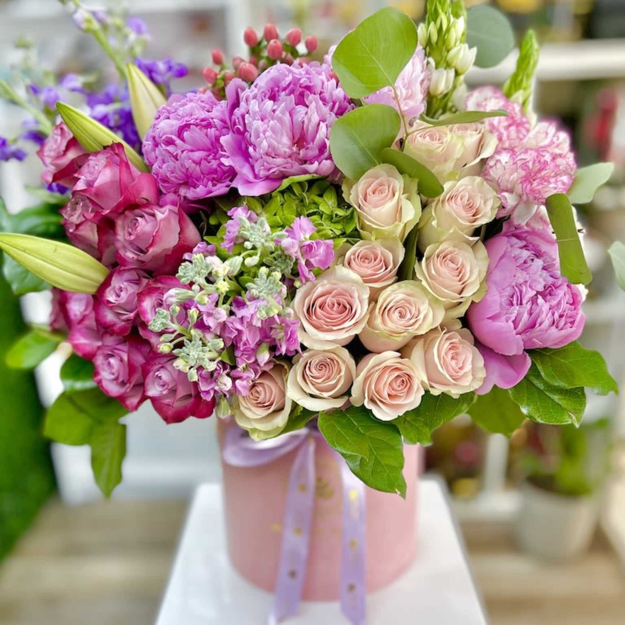 Pink and blush roses, peonies, and lilies arranged in a round pink hatbox with ribbon