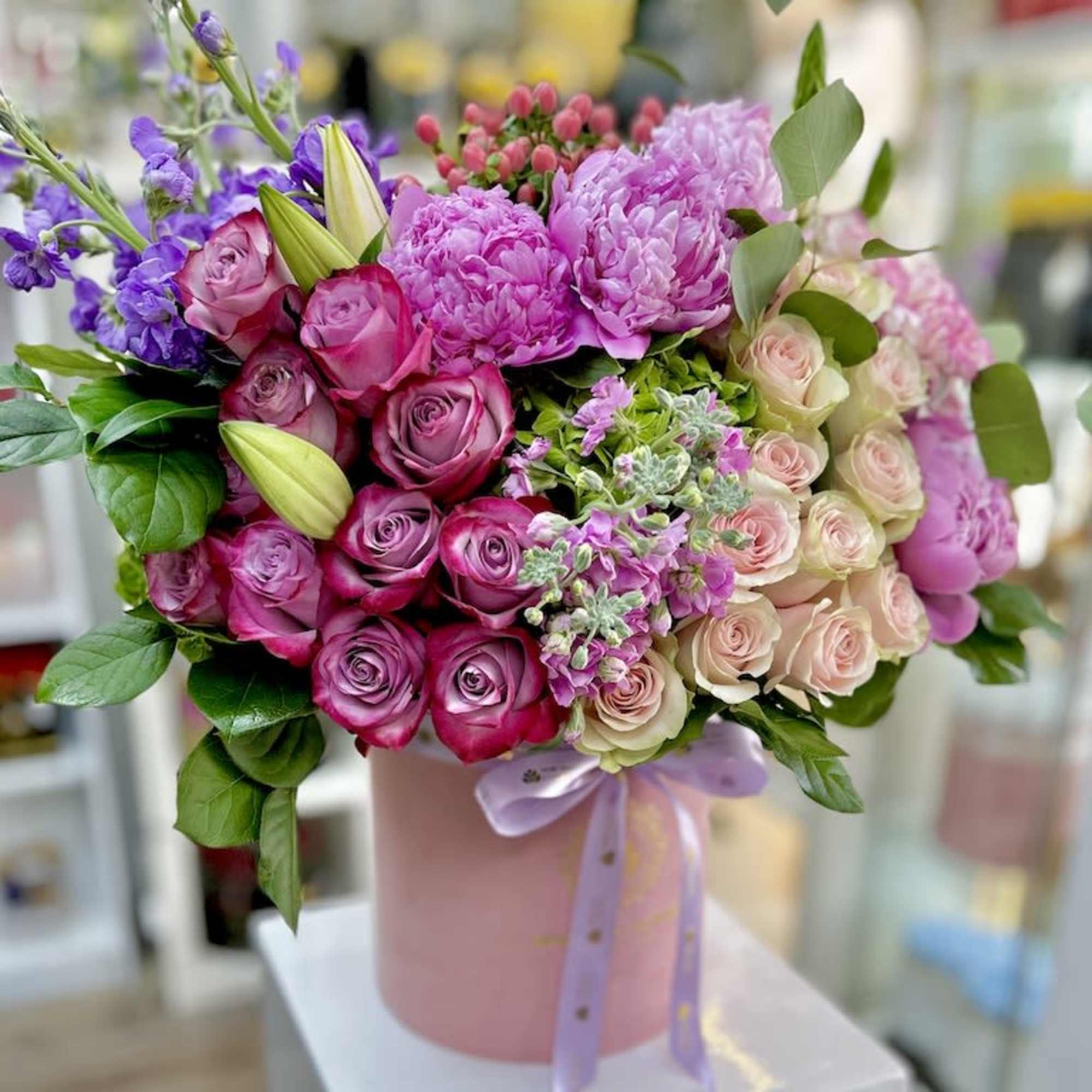 Pink hat box filled with pink roses, peonies, lilies, and purple flowers tied with a satin ribbon