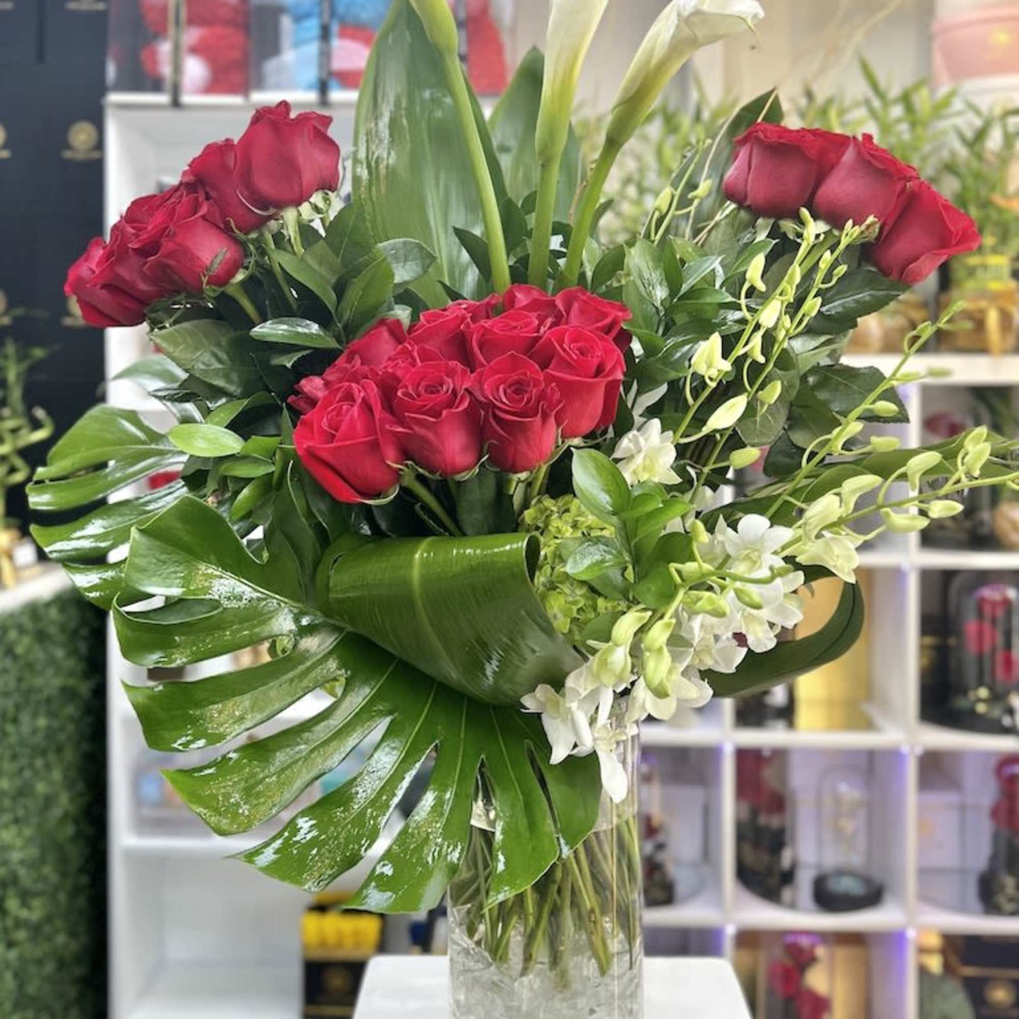 Tall arrangement of red roses, white calla lilies, and orchids in a clear glass vase with large glossy leaves