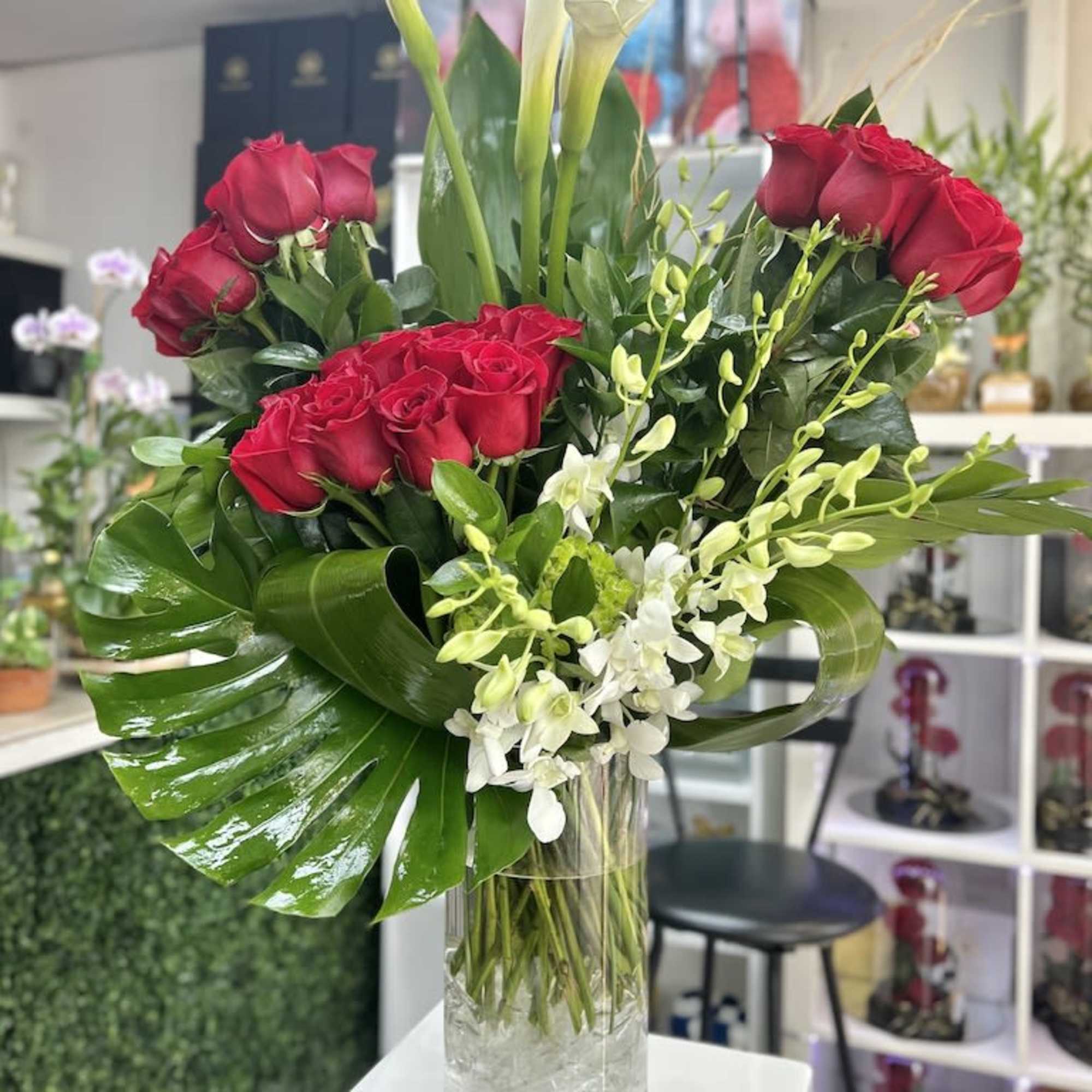 Tall arrangement of red roses, white calla lilies, orchids, and a large tropical leaf in a clear vase
