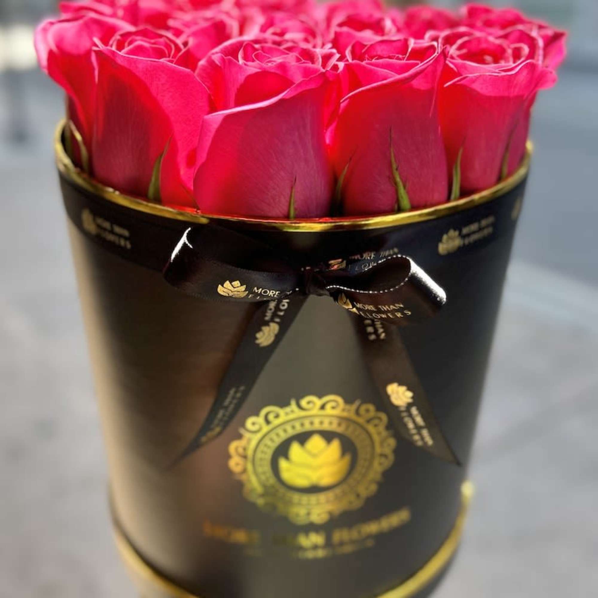 Hot pink roses arranged in a black round hat box with gold accents and ribbon