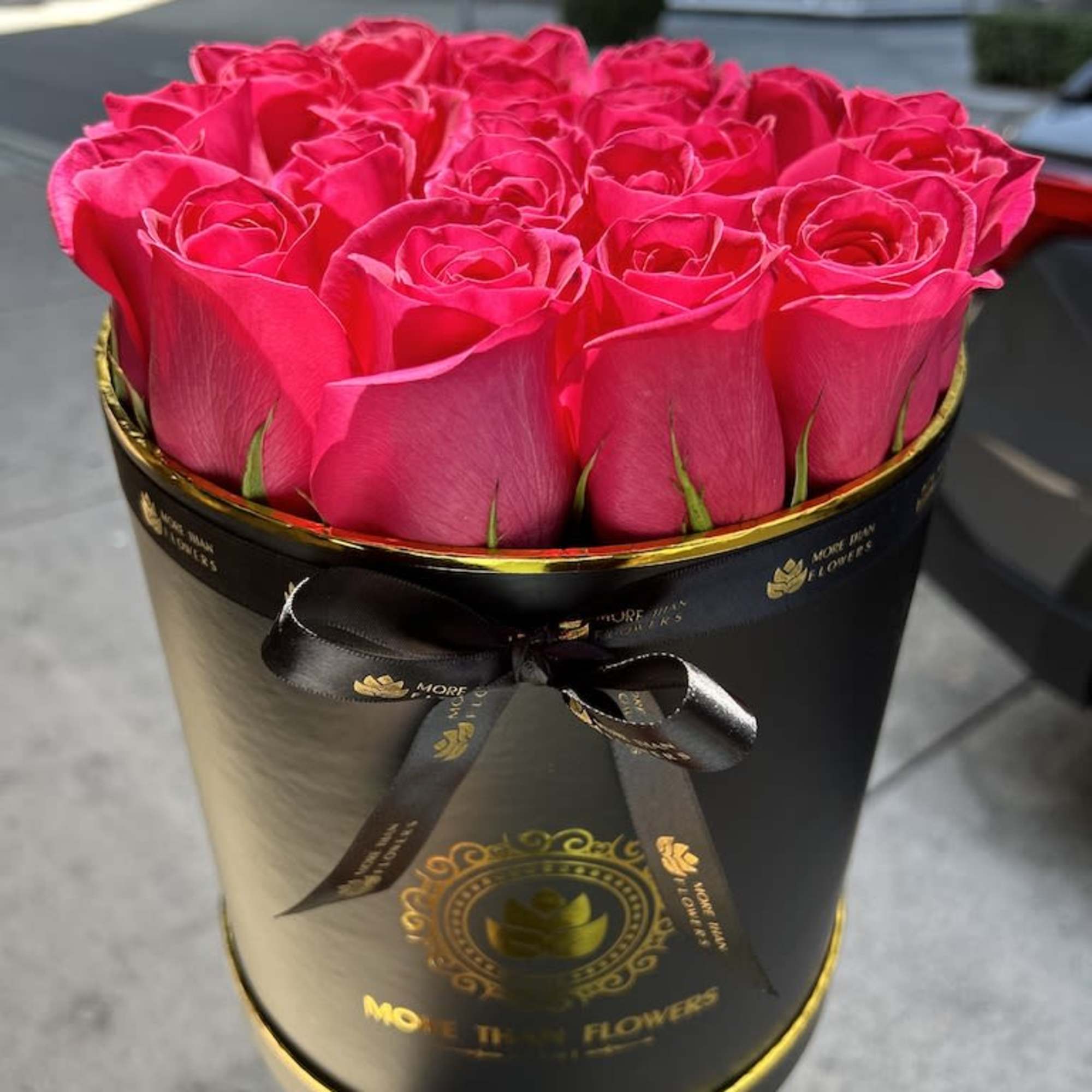 Bright pink roses arranged in a round black hatbox with gold accents and ribbon