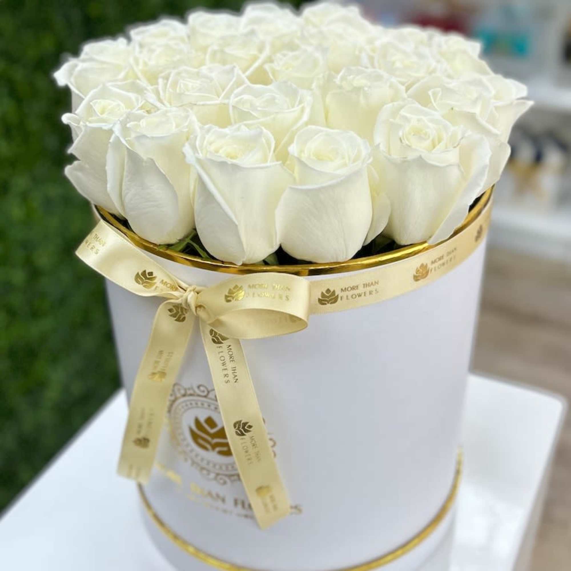 Round white hatbox filled with white roses and wrapped with a gold logo ribbon.