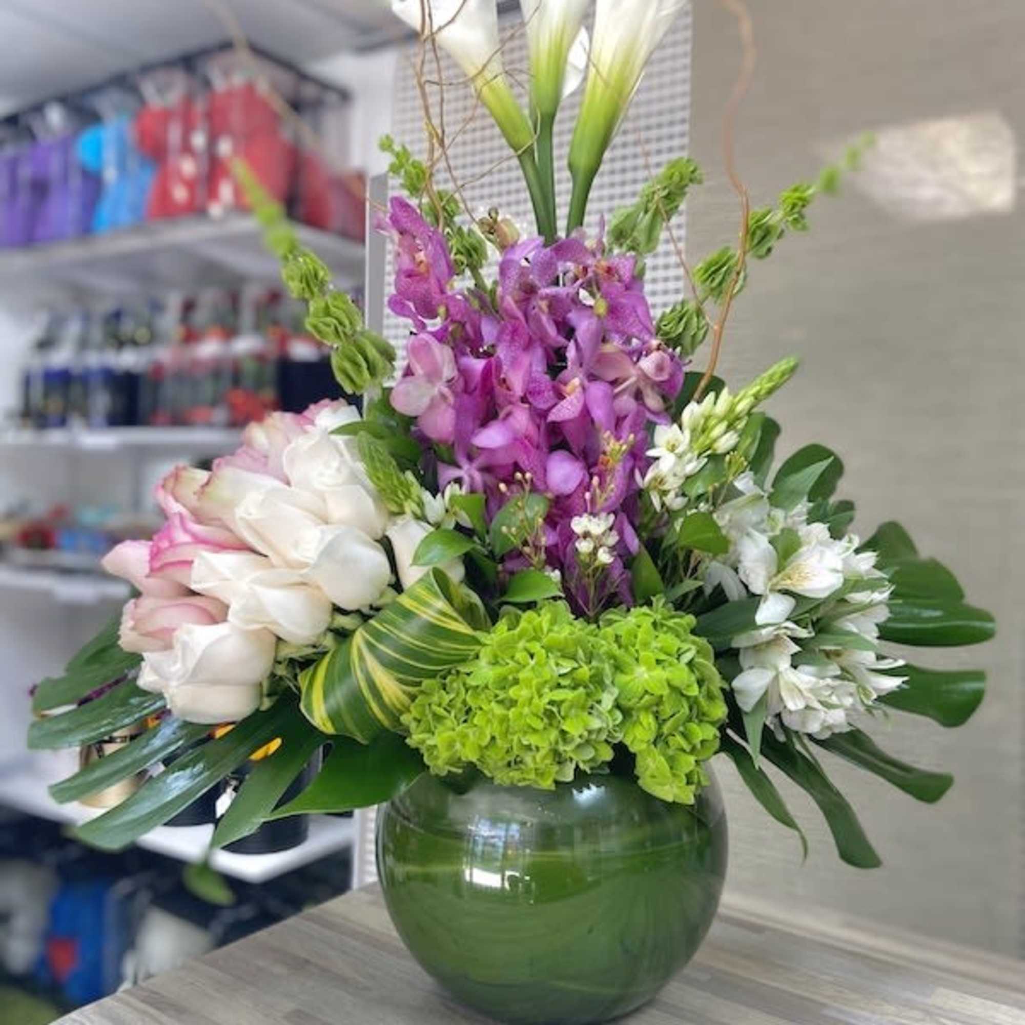 Tall arrangement of white calla lilies, roses, orchids and green hydrangea in a round green glass vase with bold leaves