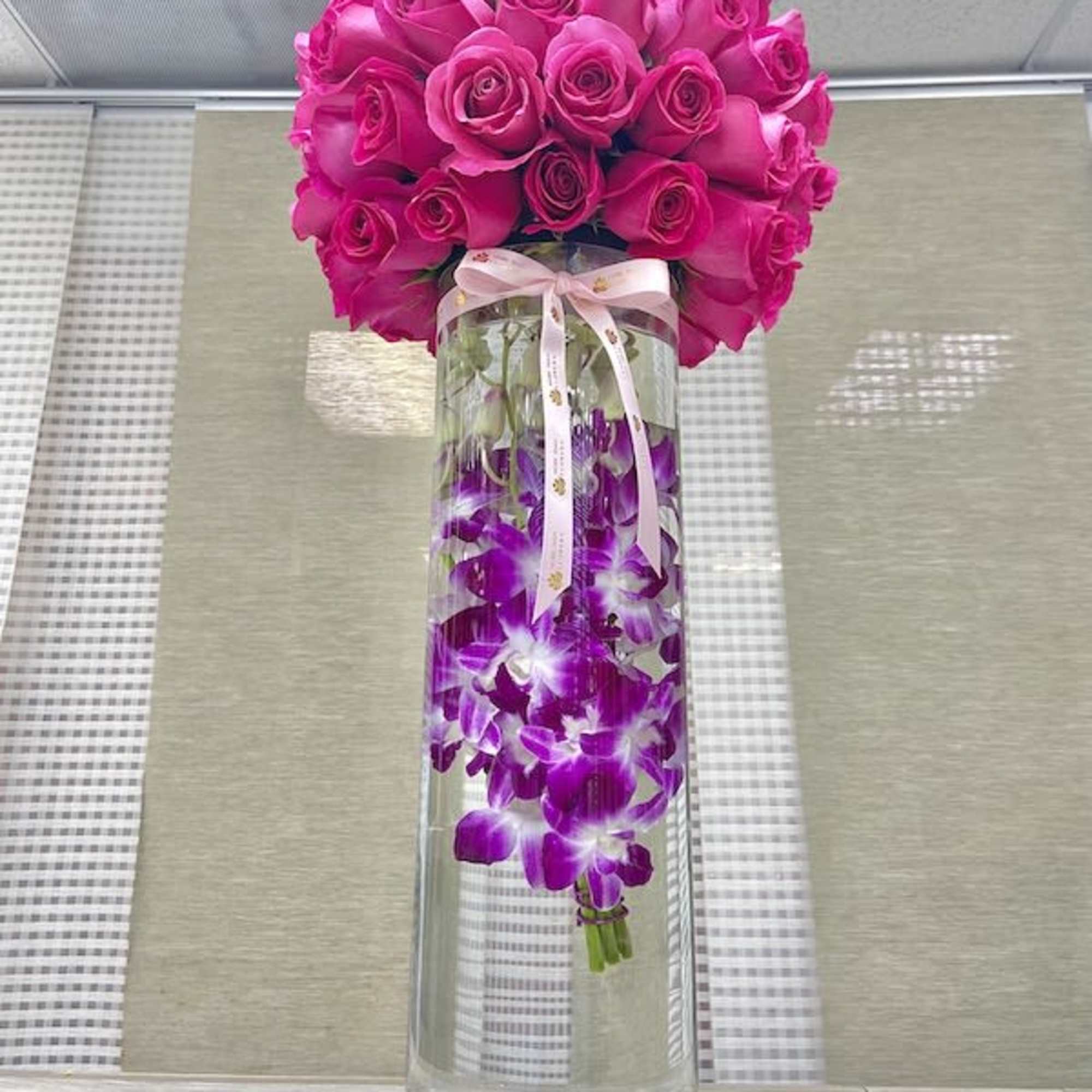 Tall glass cylinder vase with submerged purple orchids topped by dense dome of hot pink roses and a tied ribbon