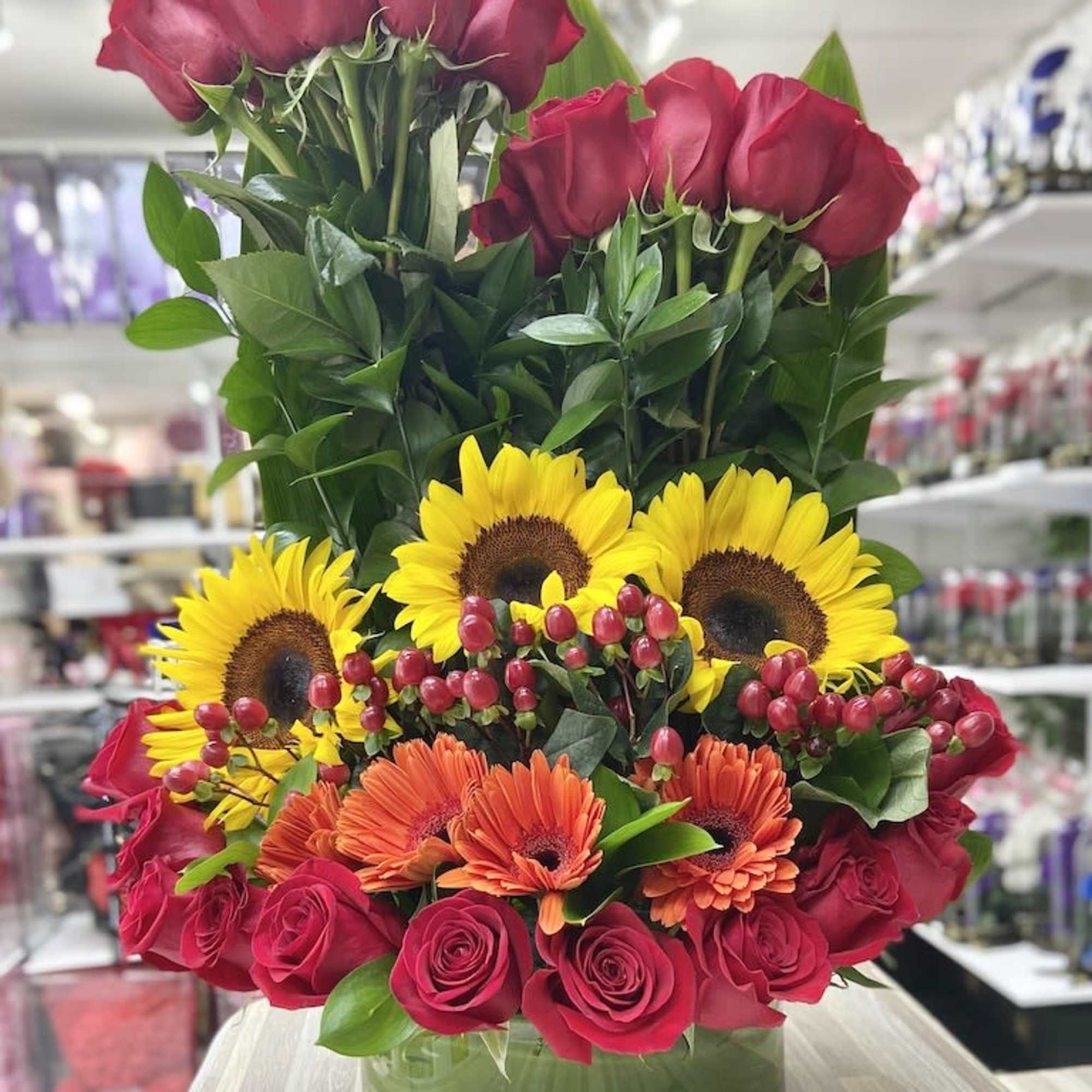 Tall arrangement of red roses, yellow sunflowers, and orange gerbera daisies in a round glass vase.