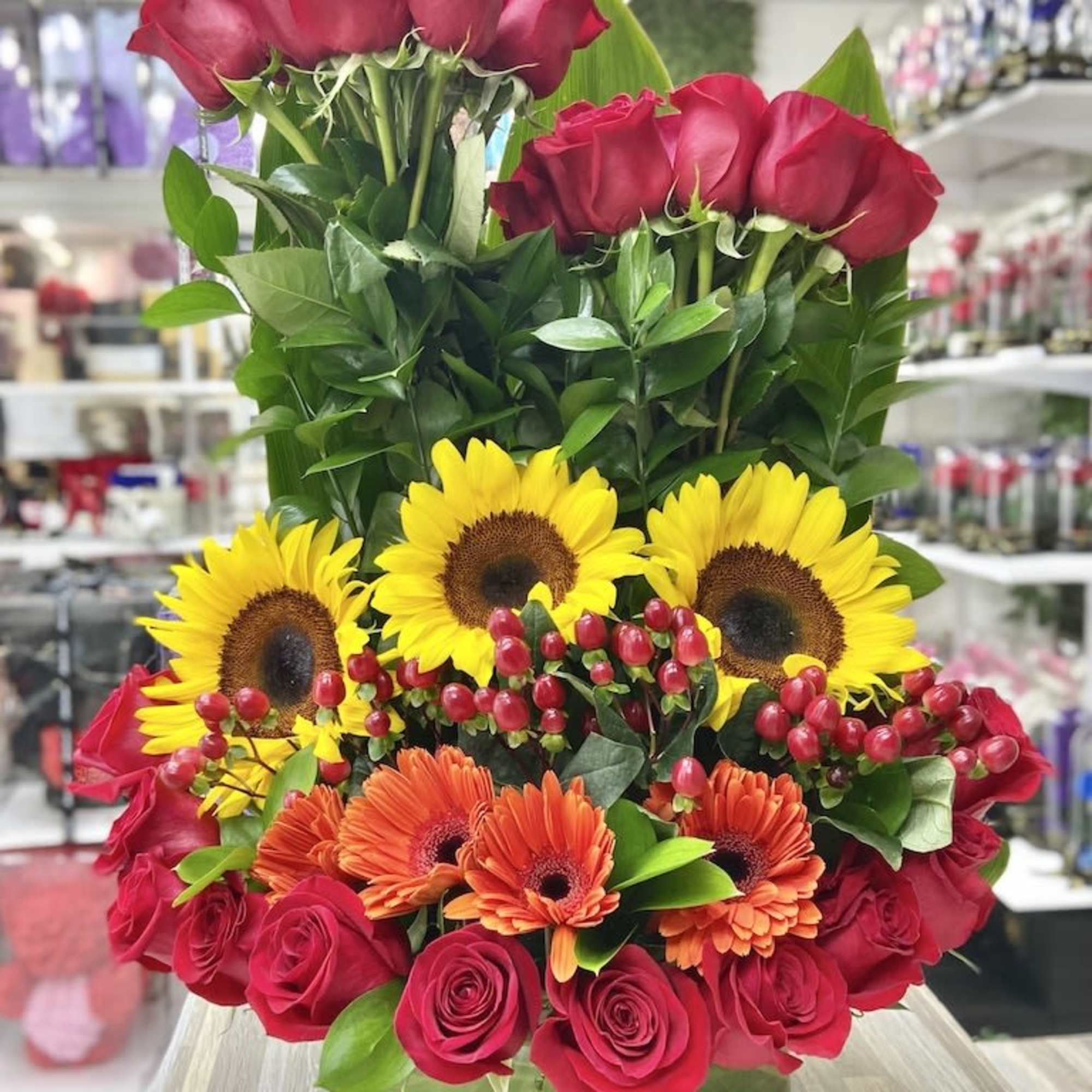 Tall arrangement of red roses, yellow sunflowers, and orange gerbera daisies in a round glass vase