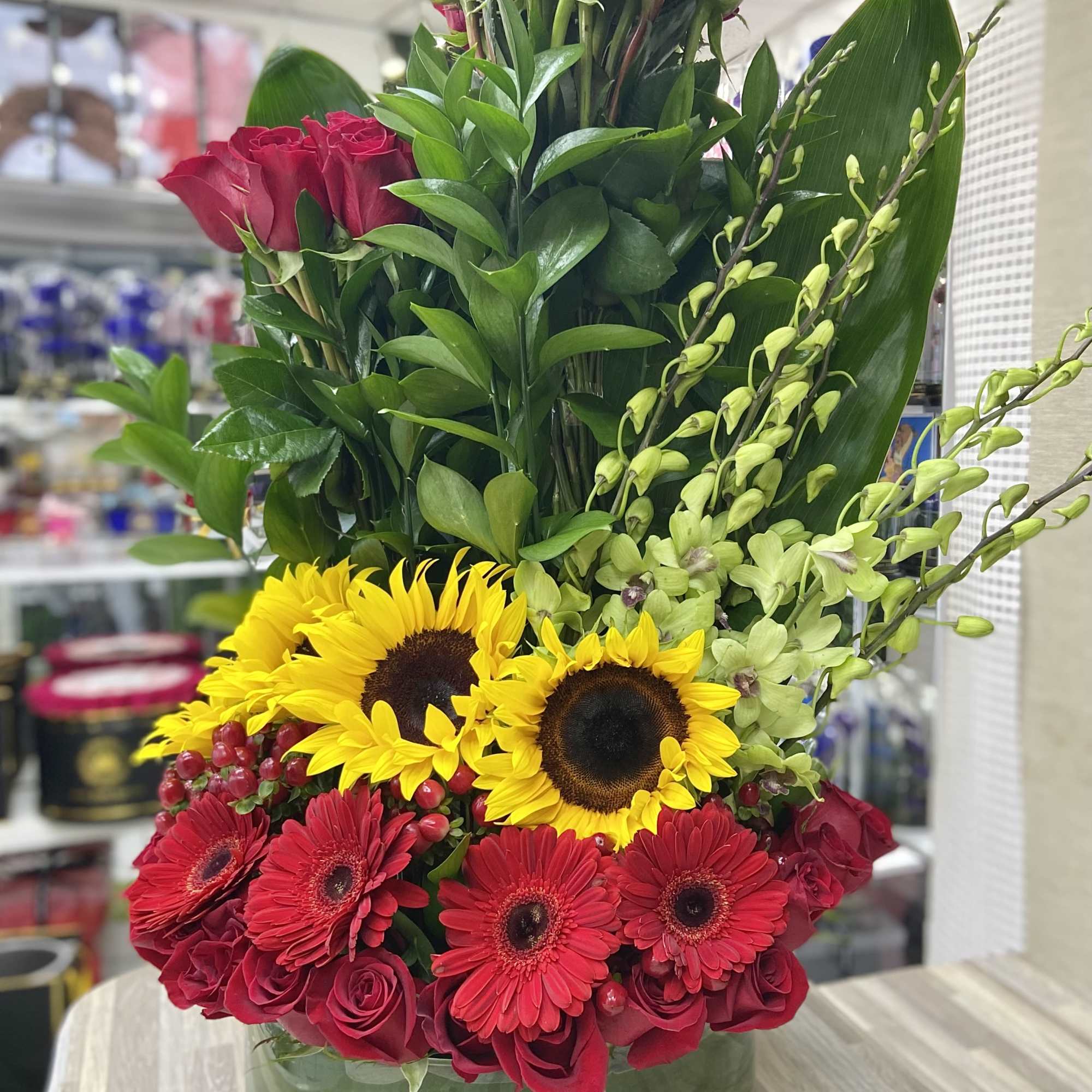 Tall arrangement of red roses, red gerberas, yellow sunflowers and green orchids in a round glass vase