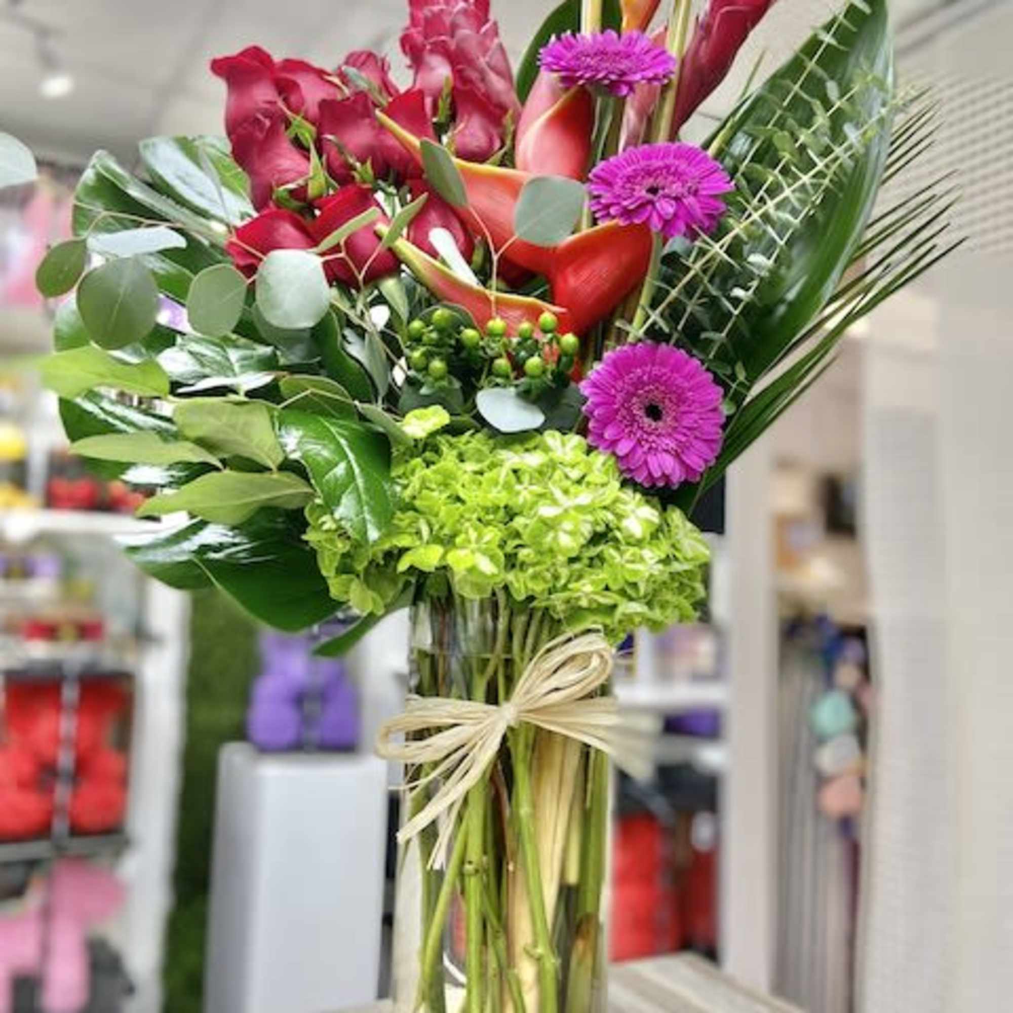 Tall tropical vase arrangement with pink gerbera, red callas, green hydrangea, and bold foliage.