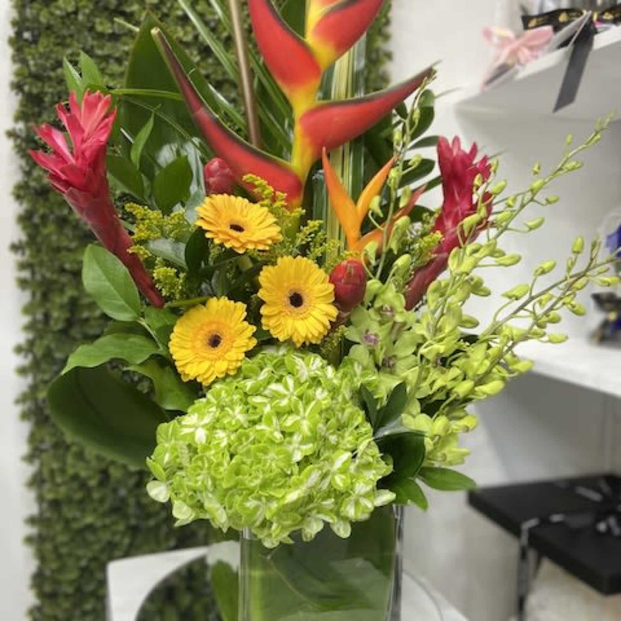 Tall tropical arrangement with red heliconia, yellow gerberas, orchids, and green hydrangea in a glass cube vase