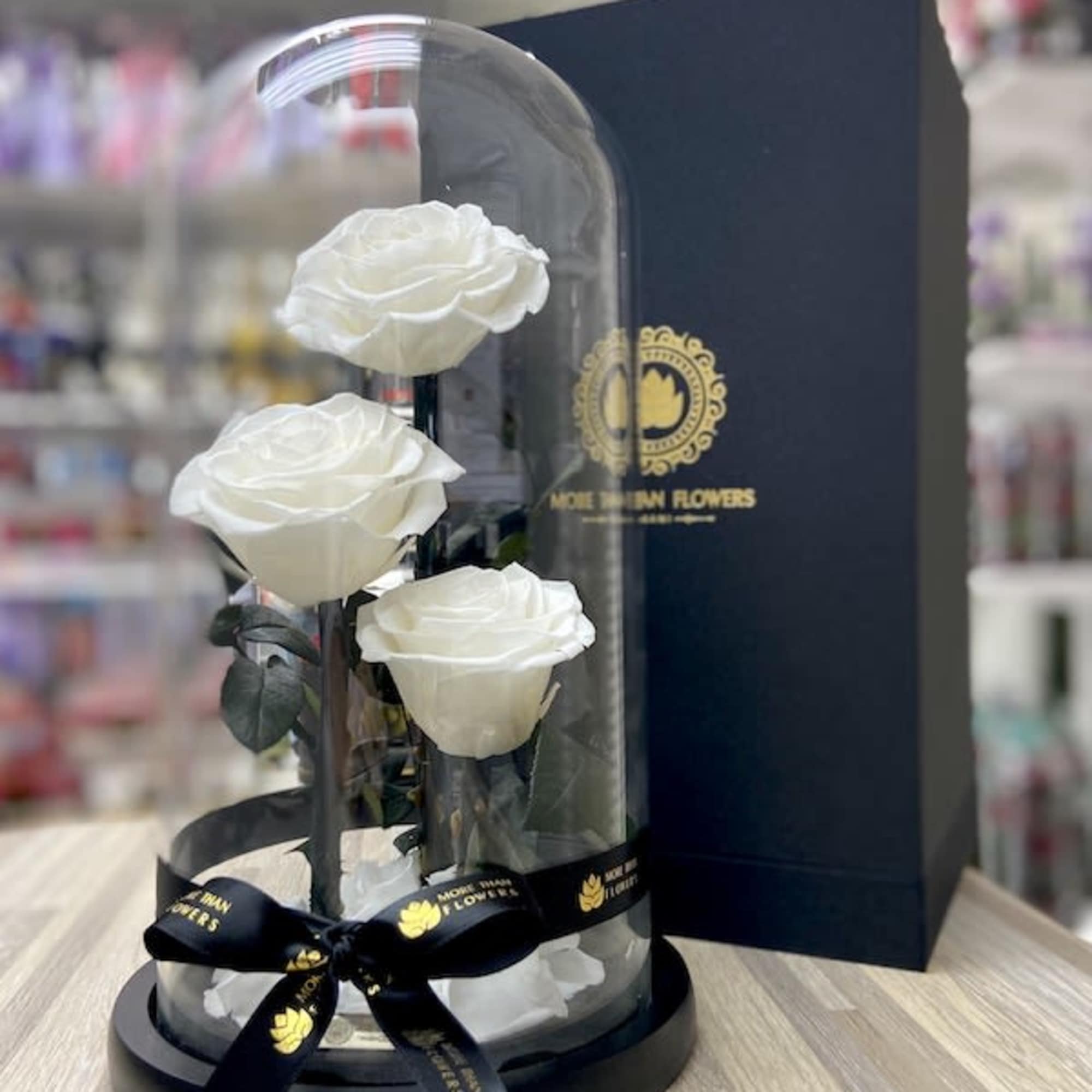 Three white roses displayed under a glass dome on a black base with a ribbon and gift box.
